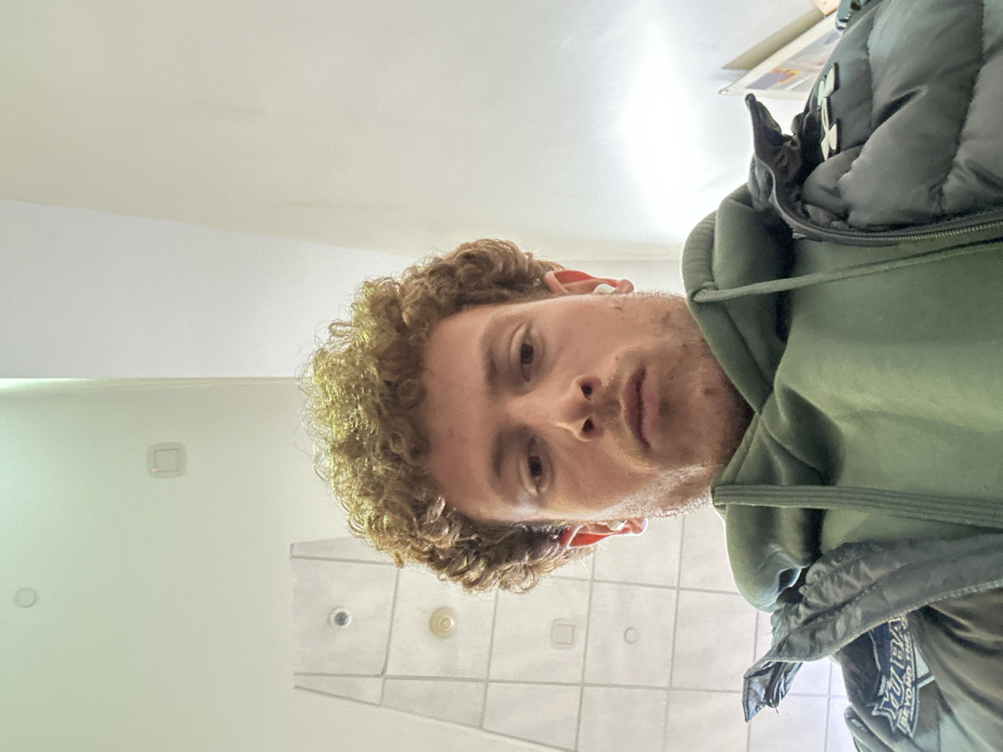 A man with curly blonde hair, wearing a green hooded sweatshirt and a puffy black jacket, is taking a selfie in what appears to be a bathroom. The background includes white walls and tiled flooring, indicating the setting is indoors. The man is looking down at the camera.
There is no indication as to whether the photo was taken in New York City.