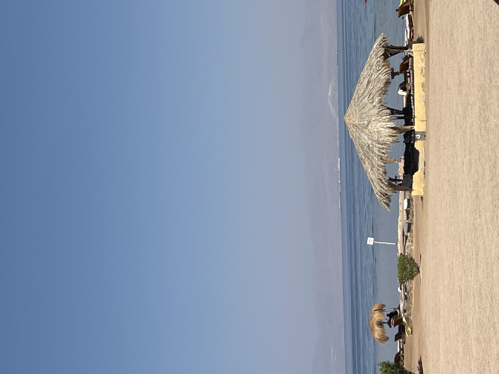 The image depicts a beach scene in Nuweiba, Egypt, under a clear blue sky. In the foreground, a sandy beach stretches towards the water. A thatched-roof hut with an open-air seating area is prominent in the center, offering shade. To the left, another thatched umbrella shades two lounge chairs, and a yellow kayak rests on the sand. The sea, a calm blue, is visible behind the beach. In the distance, a hazy mountain range forms the horizon, with several ships appearing as small white specks on the water. The overall atmosphere is one of a tranquil coastal resort.