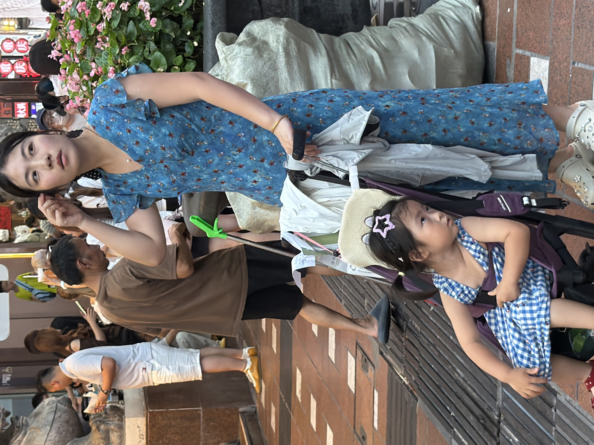 A woman in a blue floral dress is holding a stroller with a young girl inside. The girl is wearing a blue and white checkered dress and a straw hat with a star accessory. The woman is looking towards the camera with a neutral expression. Several other people are visible in the background, including a man in a brown t-shirt and a person in a white t-shirt and white shorts. The scene appears to be outdoors on a sidewalk, with brick pavement and some greenery, possibly a park or a street with shops. The lighting suggests it might be daytime, perhaps late afternoon. The overall atmosphere seems casual and busy, possibly during a shopping trip or a walk. There are no visible text elements in the provided image.