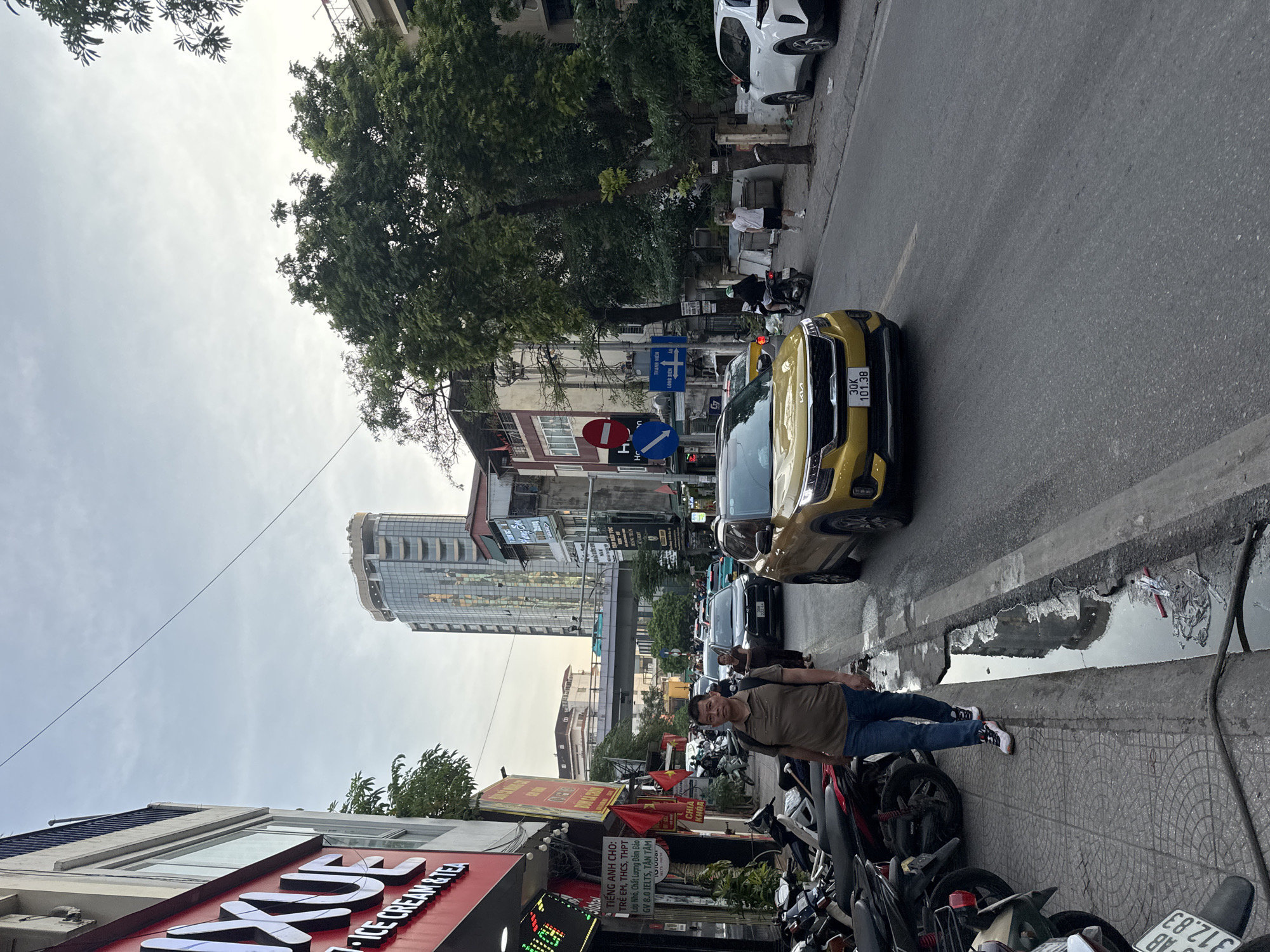 This is a vertical, eye-level, full-shot photograph taken on a street in Hanoi, Vietnam. The image captures a bustling urban scene with a focus on a man standing on the sidewalk. He is wearing a brown t-shirt, blue jeans, and white sneakers. He is looking towards the camera with a neutral expression.
To his left, a yellow Kia Seltos car is parked on the street, with its license plate visible as "30K 101.38". Behind the car, other vehicles, including motorcycles and cars, are visible, suggesting a busy traffic environment. The sidewalk is lined with parked motorcycles, a common sight in Hanoi.
In the background, there are several buildings, including a modern glass skyscraper and older, multi-story structures with signs. One prominent sign on a building reads "ICE CREAM & TEA". Other Vietnamese text is visible on signs and banners, including "TIẾNG ANH CHO: TRẺ EM, THCS, THPT" which translates to "English for: Children, Middle School, High School".
The sky is overcast, indicating a cloudy day, and there are trees with dense foliage lining the street. The overall impression is a snapshot of everyday life on a city street, with a mix of modern and traditional elements. The time of day appears to be late afternoon or early evening, given the lighting conditions.