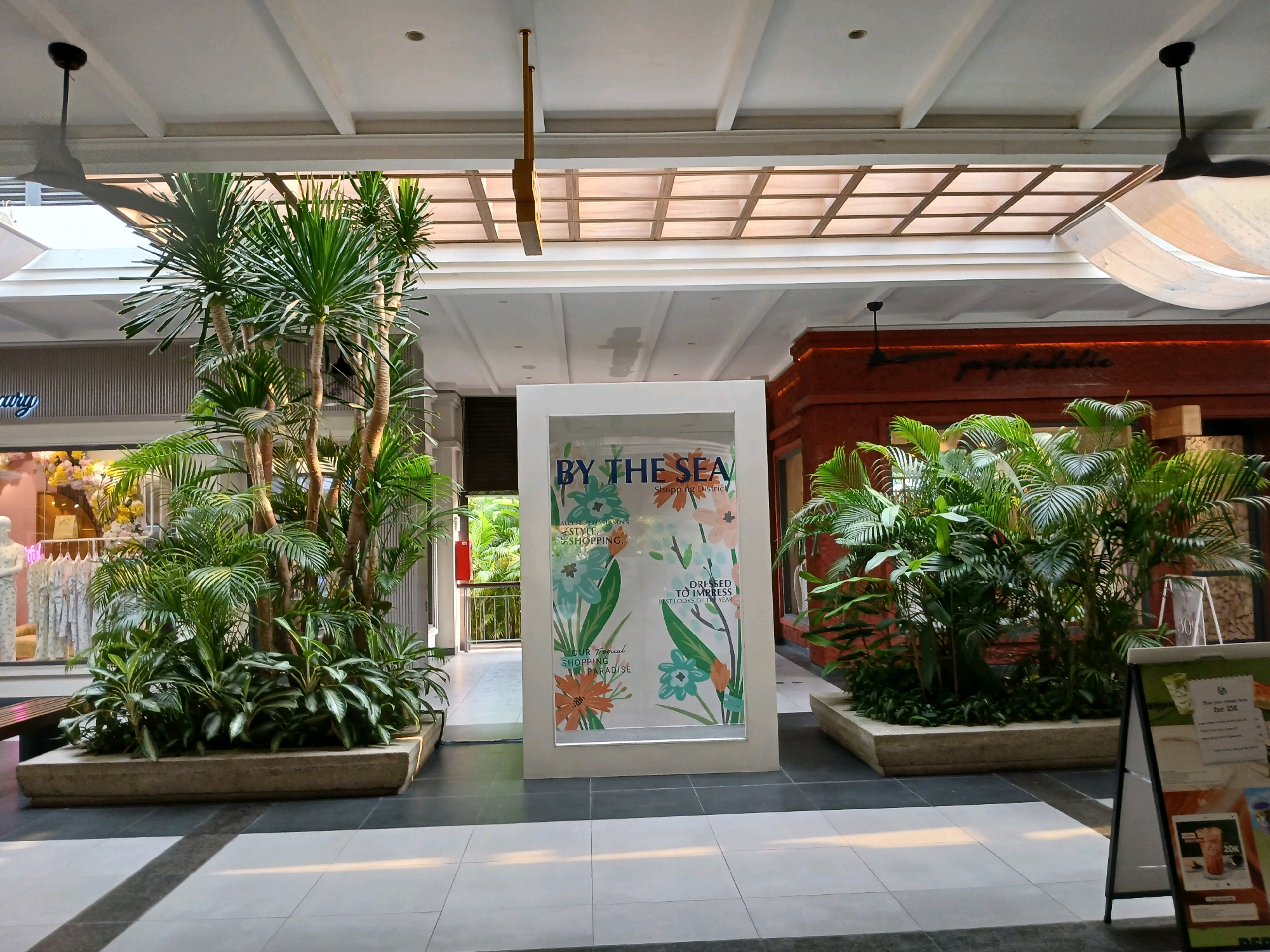 The image shows an indoor commercial area, likely a mall, in North Jakarta, Indonesia. The space features high ceilings with a grid-like pattern and exposed beams. A large display unit in the center of the frame showcases a poster with floral motifs and the text "BY THE SEA SHOPPING DISTRICT". To the left and right of the display are large planters filled with lush green tropical plants, including palm trees. In the background, to the left, are shopfronts with mannequins and clothing displays. To the right, there is a glimpse of another shop with a reddish-brown facade. The floor is tiled in a checkerboard pattern of light and dark gray squares. There are ceiling fans visible, one on the left casting a shadow. An A-frame sign is partially visible in the lower right corner, displaying some promotional information. The overall impression is of a well-lit, air-conditioned commercial environment designed with tropical aesthetics.