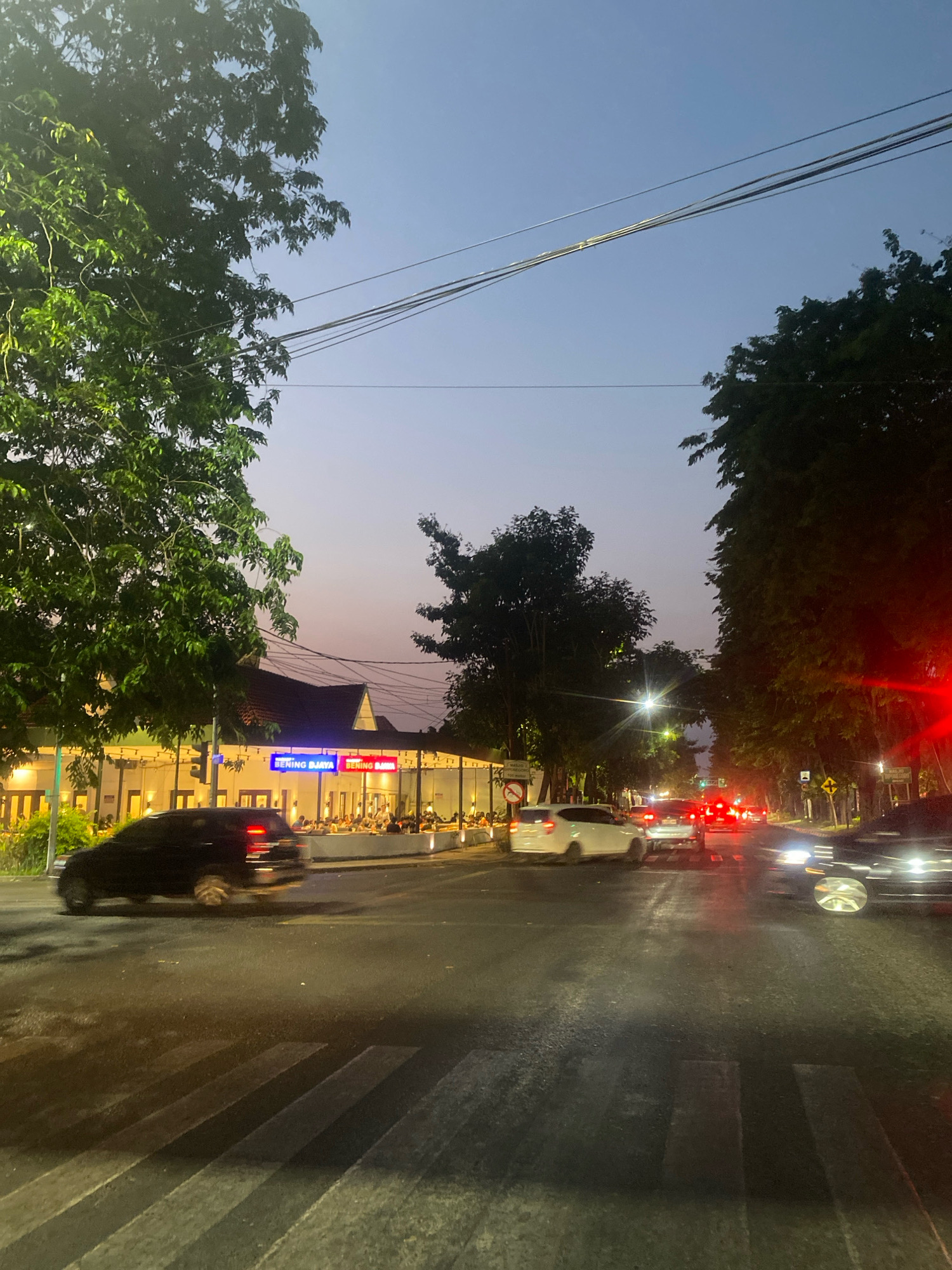 The image depicts a street scene at dusk in an urban environment. Several cars are present on the wet asphalt road, with some headlights and taillights illuminated. A black SUV is visible in the foreground, captured mid-motion as it crosses a pedestrian crossing. Further down the street, other vehicles are also in motion or stopped, their lights creating streaks of color against the darkening sky.
On the left side of the street, a building with a tiled roof is illuminated by warm interior lights. Signs with blue and red lettering are visible on the building's facade. Many people are seated at tables outside the building, suggesting it is a restaurant or dining establishment. Lush green trees line the sides of the street, their leaves casting shadows on the road. Overhead, power lines stretch across the sky. The sky itself is a gradient of pale blue and purple, indicating the transition from day to night.