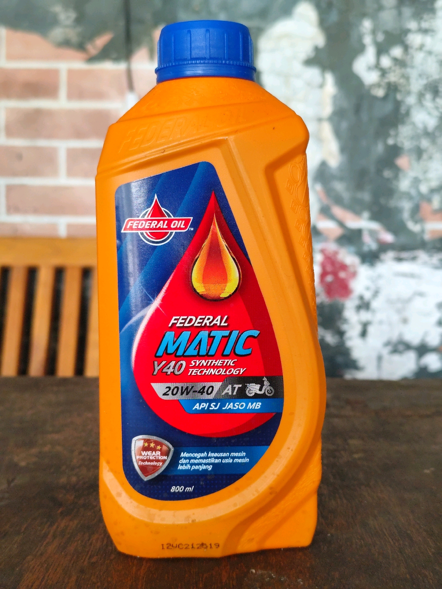 An orange plastic bottle of Federal Oil Matic 20W-40 synthetic motorcycle oil with a blue cap is positioned on a wooden surface. The label features a red and blue design with a depiction of a flame-like oil drop. Text on the label indicates "FEDERAL OIL", "MATIC", "SYNTHETIC TECHNOLOGY", "20W-40 AT", and "API SJ JASO MB". There is also a shield icon with "WEAR PROTECTION Technology" and text in Indonesian that translates to "Prevents engine wear and ensures longer engine life". The volume is stated as "800 ml". The background is blurred, showing a brick wall pattern and a lighter, textured surface. A portion of a wooden chair is visible to the left of the bottle. A date stamp "12VC212019" is visible on the bottom front of the bottle. The image was captured in Palangka Raya, Indonesia.