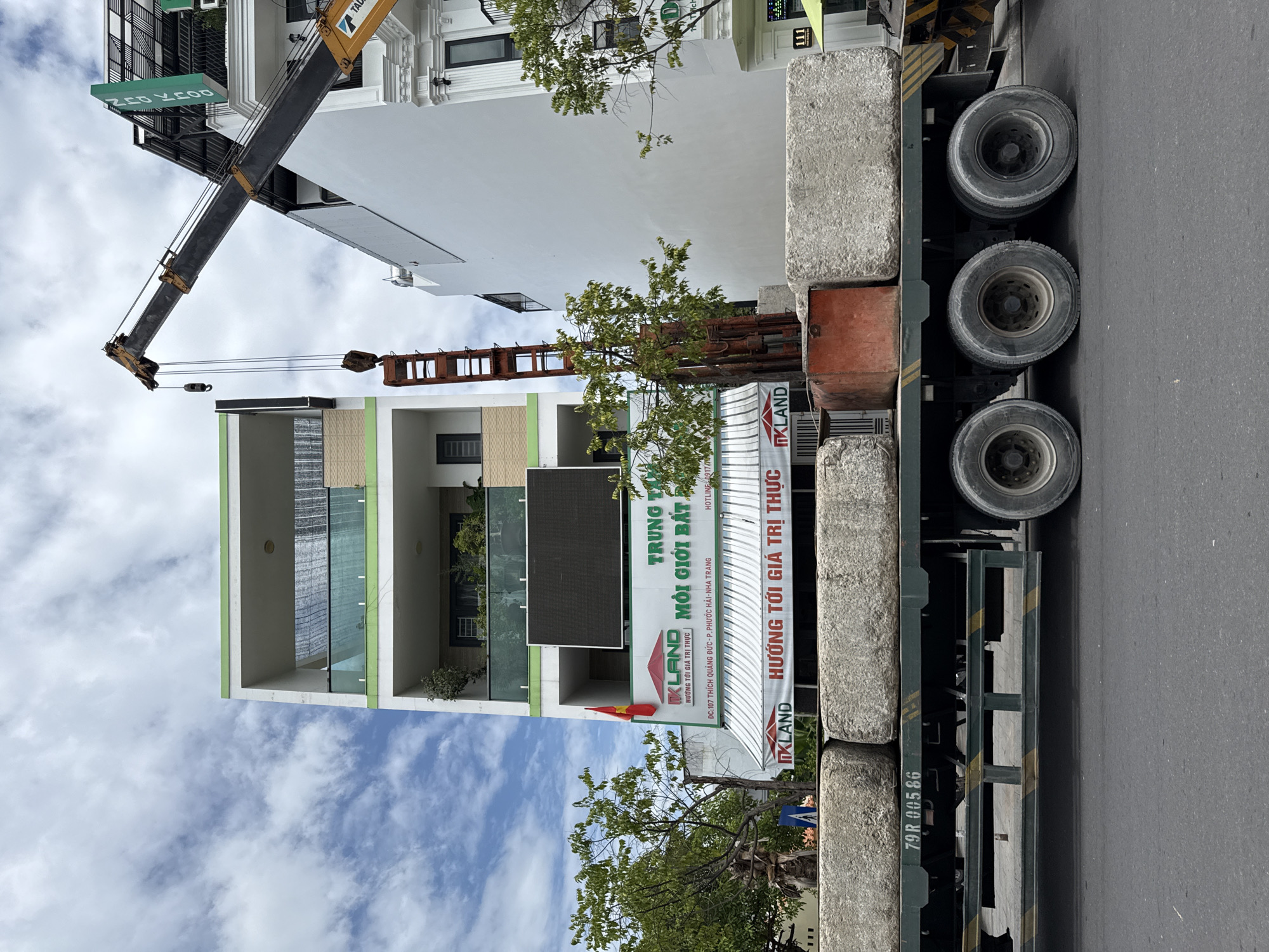 The image shows a large truck with four wheels in the foreground, loaded with several large, rectangular concrete blocks. The truck is parked on a street in Nha Trang, Vietnam. Behind the truck, a building with a white facade and green trim is visible, with several balconies. A large LED screen is mounted on the building. A crane is extending over the building, with a load suspended from it. Several trees are also present in the scene. The sky is cloudy. The building appears to be a commercial establishment, with signs in Vietnamese indicating "IKLAND" and "MÔI GIỚI BẤT ĐỘNG SẢN," which translates to "real estate broker." The address "ĐC: 107 THÍCH QUẢNG ĐỨC - P. PHƯỚC HẢI - NHA TRANG" is also visible on a sign, confirming the location as Nha Trang. The scene depicts a construction or delivery operation.