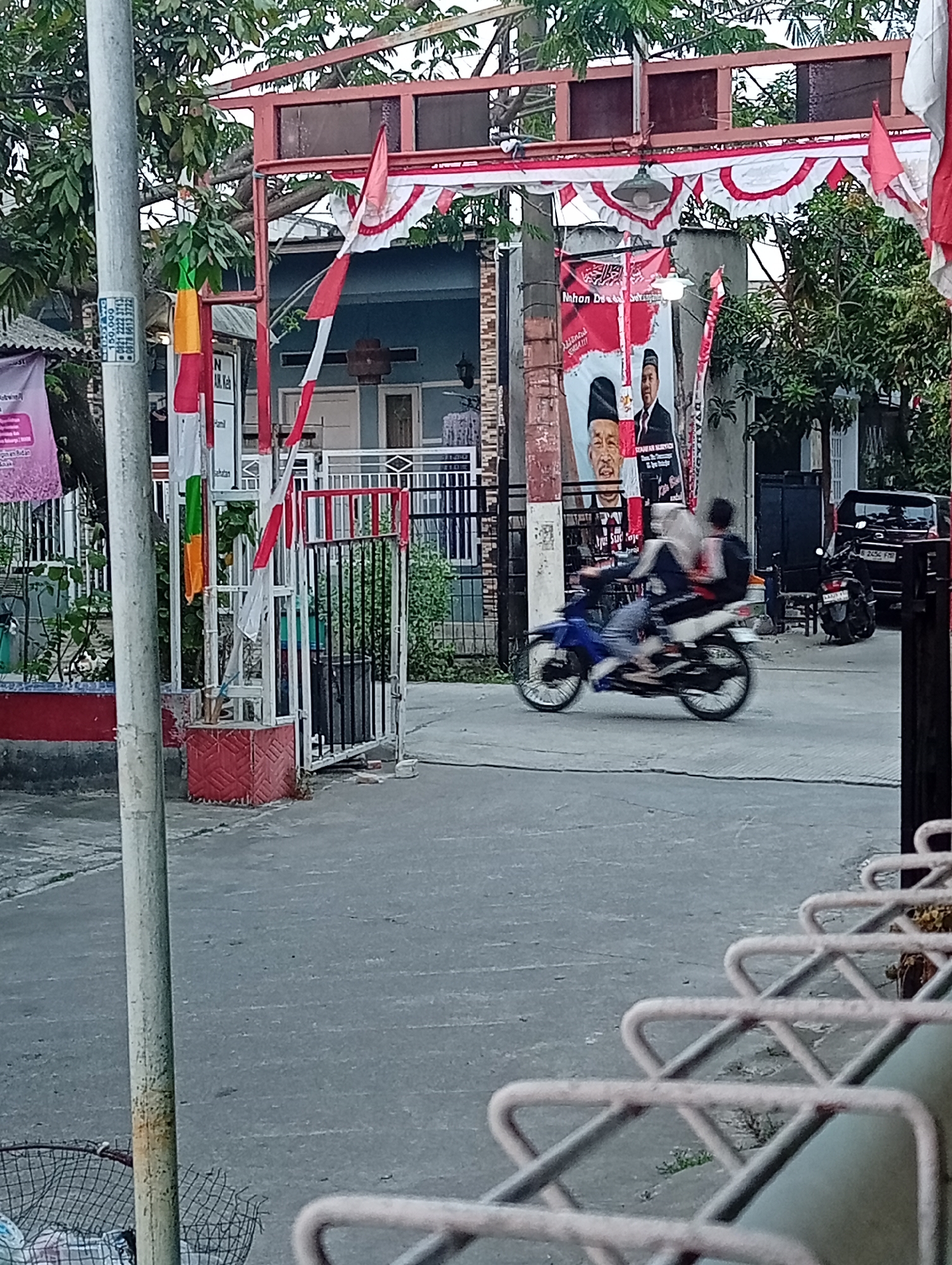 In Wanajaya, Indonesia, this image captures a street scene featuring a white metal gate adorned with red and white Indonesian flags. A large poster with images of two men and text is visible behind the gate, with red and white bunting stretched across the top of the scene. A blue motorcycle with two blurred figures, one driving and one as a passenger, moves from left to right across the foreground. A dark-colored SUV is parked to the right of the motorcycle, and another motorcycle is parked behind it. A concrete pole stands on the left side of the image, and a metal trash bin is located at its base. The ground is paved with asphalt, and various green trees and bushes are present in the background. The overall impression is one of a typical day in a residential or semi-commercial area of the city, possibly during a celebratory period given the flags and decorations.