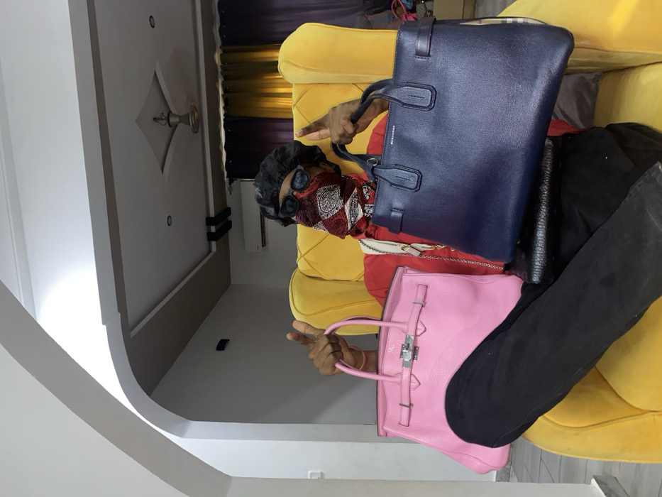 This image depicts an individual seated in a yellow upholstered armchair within a modern interior in Tema, Ghana. The person is wearing a black durag, dark sunglasses, a red, white, and black patterned bandana covering the lower face, a red long-sleeved shirt, and black pants. Both hands are raised, each forming a thumbs-up gesture while holding a handbag. The left hand holds a pink textured leather handbag with prominent silver hardware, including a lock, clasp, and buckles. The right hand holds a dark blue textured leather tote bag, visibly branded "BURBERRY." A black cross-body bag is slung across the individual. The background features white walls, including a curved archway on the left. The ceiling is a multi-tiered false design with recessed lighting and a central fixture. A wall-mounted air conditioning unit is visible on the back wall. Dark curtains hang on the right side of the frame, and a partially visible patterned bag is discernible near the curtains behind the blue tote.