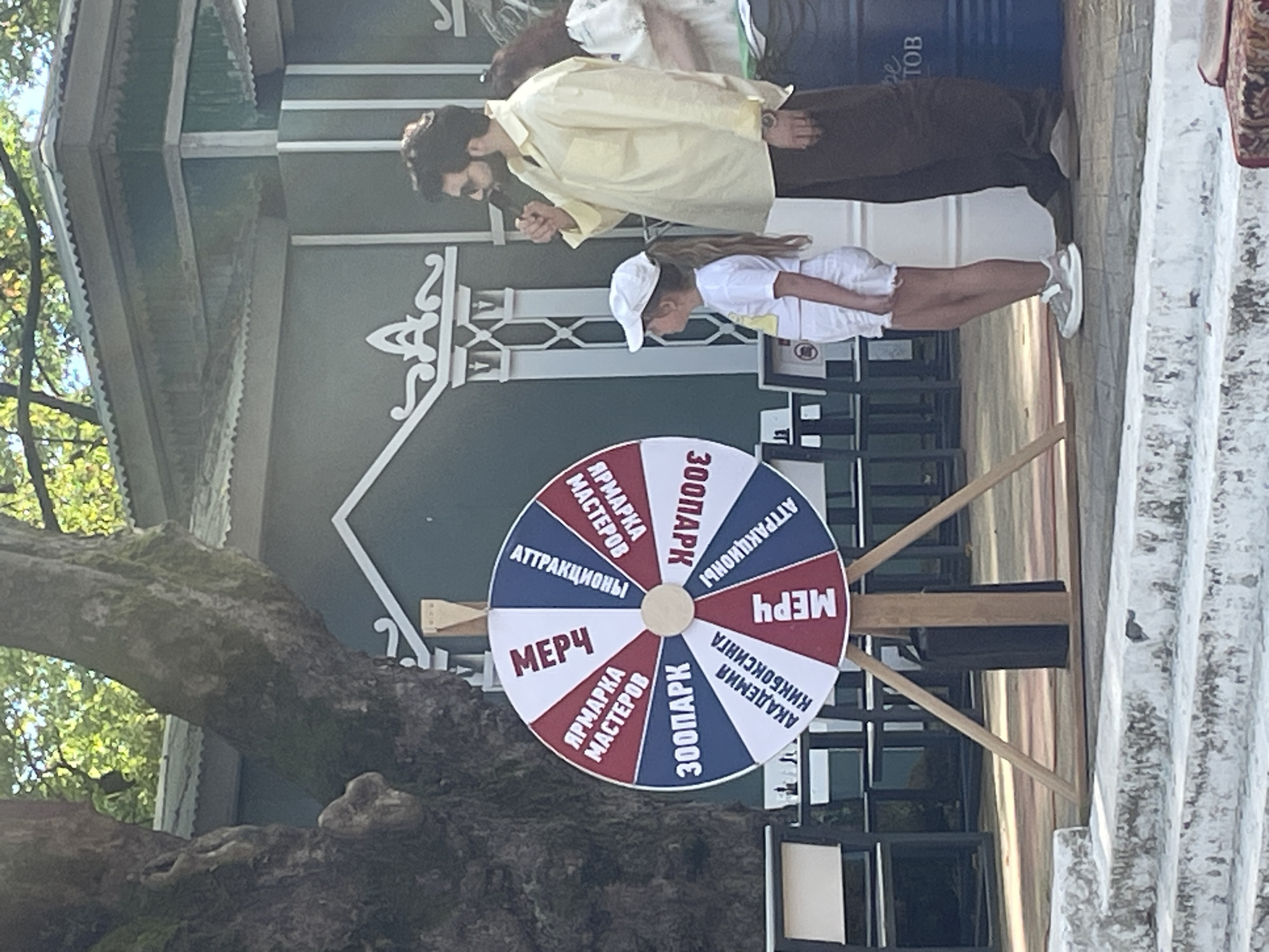 This image depicts a lively outdoor scene, likely in Sochi, Russia, centered around a prize wheel. A man, dressed in a light yellow shirt and dark trousers, stands to the right, holding a microphone and speaking. Beside him, a young girl in a white cap, t-shirt, and shorts stands attentively, facing the wheel. The prize wheel itself is a prominent feature, divided into red, white, and blue sections with various text inscriptions. Some visible inscriptions on the wheel include "МЕРЧ" (Merch), "ЯРМАРКА МАСТЕРОВ" (Craft Fair), "300ПАРК" (300Park), and "АТТРАКЦИОНЫ" (Attractions), and "АКАДЕМИЯ КИКБОКСИНГА" (Kickboxing Academy). The wheel is mounted on a wooden stand with three legs. In the background, a large, old tree with moss-covered bark stands to the left. Behind the wheel and people, there's a building with green walls and decorative white trim, and a corrugated metal roof. The ground appears to be paved, with a set of concrete steps in the foreground. The overall atmosphere suggests an event or attraction, possibly at a park or fair.
