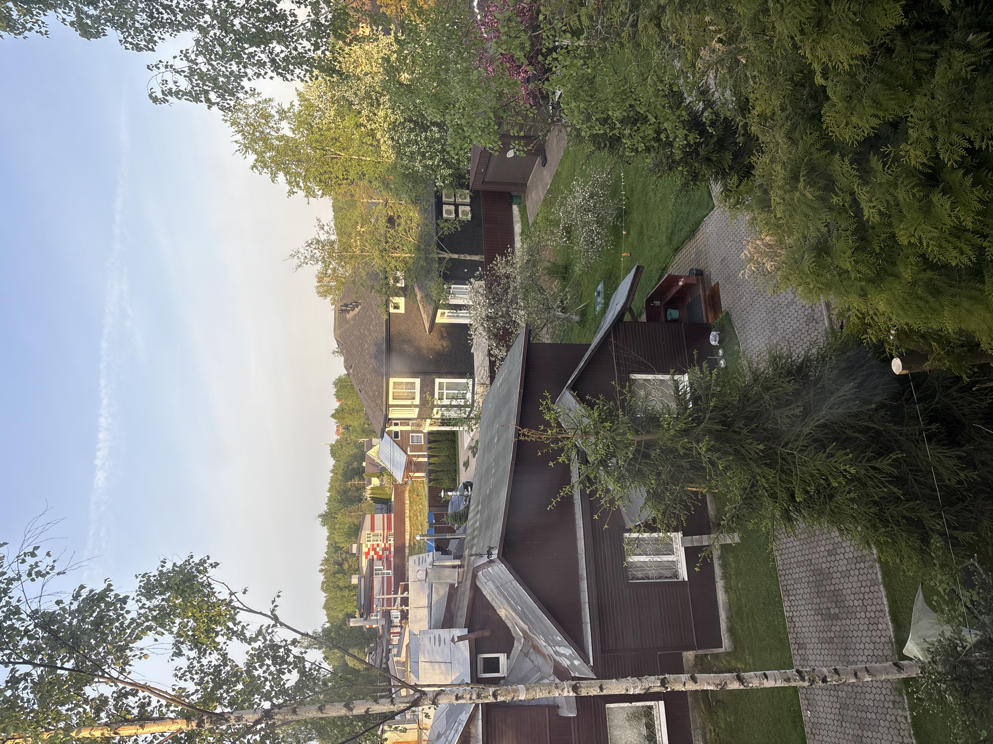 The image depicts a residential area in Saint Petersburg, Russia. Several wooden houses with brown and gray roofs are visible, surrounded by trees and greenery. A paved path runs alongside one of the houses. The sky is light blue. The houses are primarily one-story. Some of the houses have solar panels on their roofs. The weather appears to be sunny.