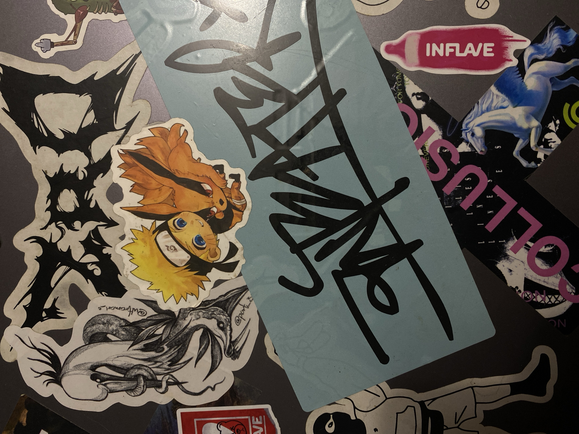 The image is a top-down view of a surface covered in various stickers. The stickers depict a range of imagery, including cartoon characters, abstract designs, graffiti-style lettering, and a horse with wings. One prominent sticker features a light blue background with black graffiti lettering. Other stickers show a black and white drawing of a creature with a snake wrapped around it, a cartoon character with orange spiky hair and a fox-like tail, a black silhouette of a person wearing a hoodie and balaclava, and a pink sticker shaped like a baby bottle with the word "INFLAVE" written vertically in white. The overall impression is one of a collection of decals applied to a flat surface, likely a laptop or similar item. There are no discernable elements that indicate a specific city or location within the image itself.