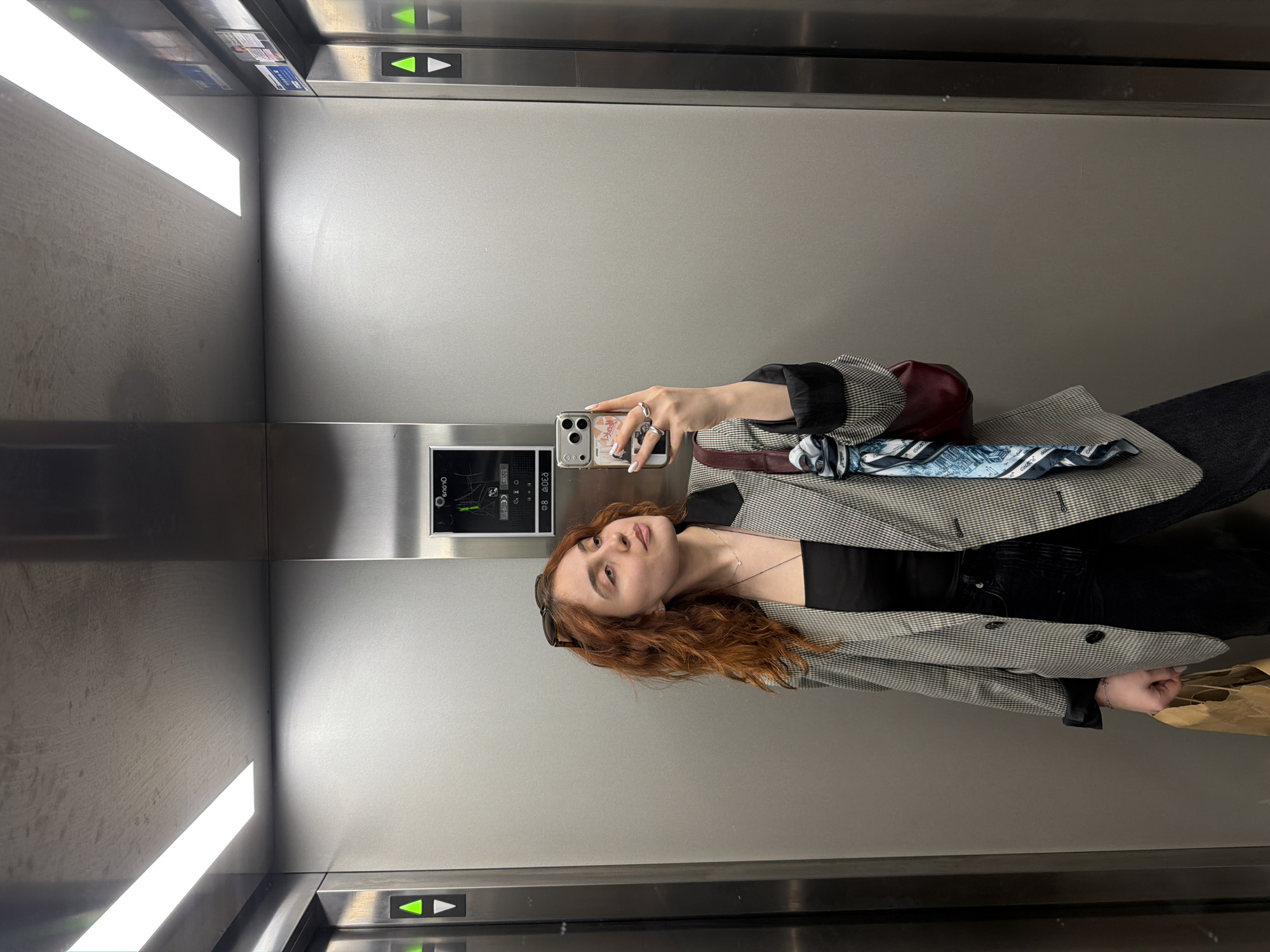 A young woman is depicted taking a selfie in what appears to be an elevator, with the image captured in a portrait orientation (rotated 90 degrees clockwise from the original view). She has reddish-brown, wavy hair, and is wearing sunglasses on her head, a black top, a black and white checkered blazer (houndstooth or similar pattern), and dark pants. Her left hand holds a smartphone with a decorated case, displaying white-tipped fingernails and rings on her fingers. A small red/maroon handbag and a blue and white patterned scarf are visible over her blazer on her right side. Part of a brown paper bag is visible near her left hand towards the bottom of the frame.
The elevator interior features light gray walls, possibly brushed metal or a laminate finish. Bright linear lights are visible on the ceiling, reflecting on the walls. The elevator controls are made of brushed stainless steel. Two illuminated arrow buttons, a green upward arrow and a white downward arrow, are prominently visible. A small digital display screen is also visible, showing a stylized "S" followed by "0100" or similar characters, and other faint diagrammatic elements, suggesting it might indicate the floor or status. The context provided indicates this scene is located in Strasbourg, France.