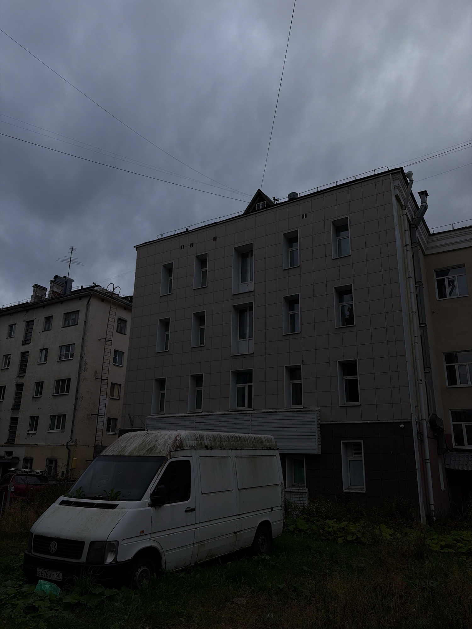 An overcast, gloomy day in Arkhangelsk, Russia, as depicted in this low-angle shot. Two apartment buildings dominate the background. The building on the right is modern, clad in beige square tiles with numerous rectangular windows. The building on the left, older and weathered, shows signs of disrepair, with multiple windows and an external fire escape ladder. In the foreground, a dilapidated white van is parked amidst overgrown grass and weeds. The van appears abandoned, its roof covered in moss and debris. The sky is a uniform, dark gray, contributing to the somber atmosphere. A few power lines stretch across the sky, and a single street light is visible near the older building. The overall scene conveys a sense of neglect and decay.