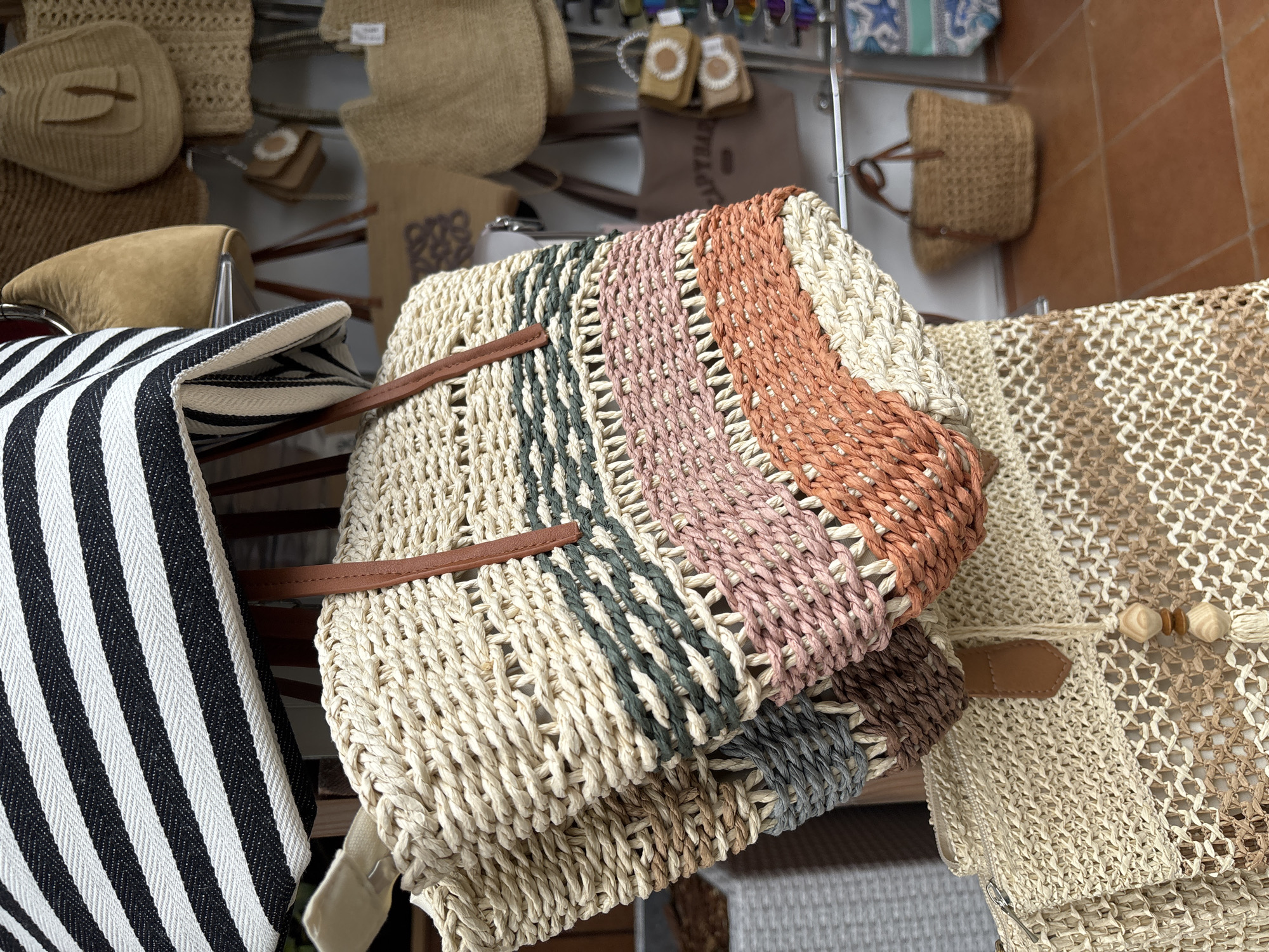 The image shows a display of woven bags, likely in a market or boutique. The bags are made of natural materials like straw or raffia and feature various weaving patterns and colors. Some bags have leather handles, and one bag in the foreground has horizontal stripes of green, pink, and orange. In the background, other bags are visible, along with shelving and what appears to be a tiled floor. The location is identified as Sochi, Russia.