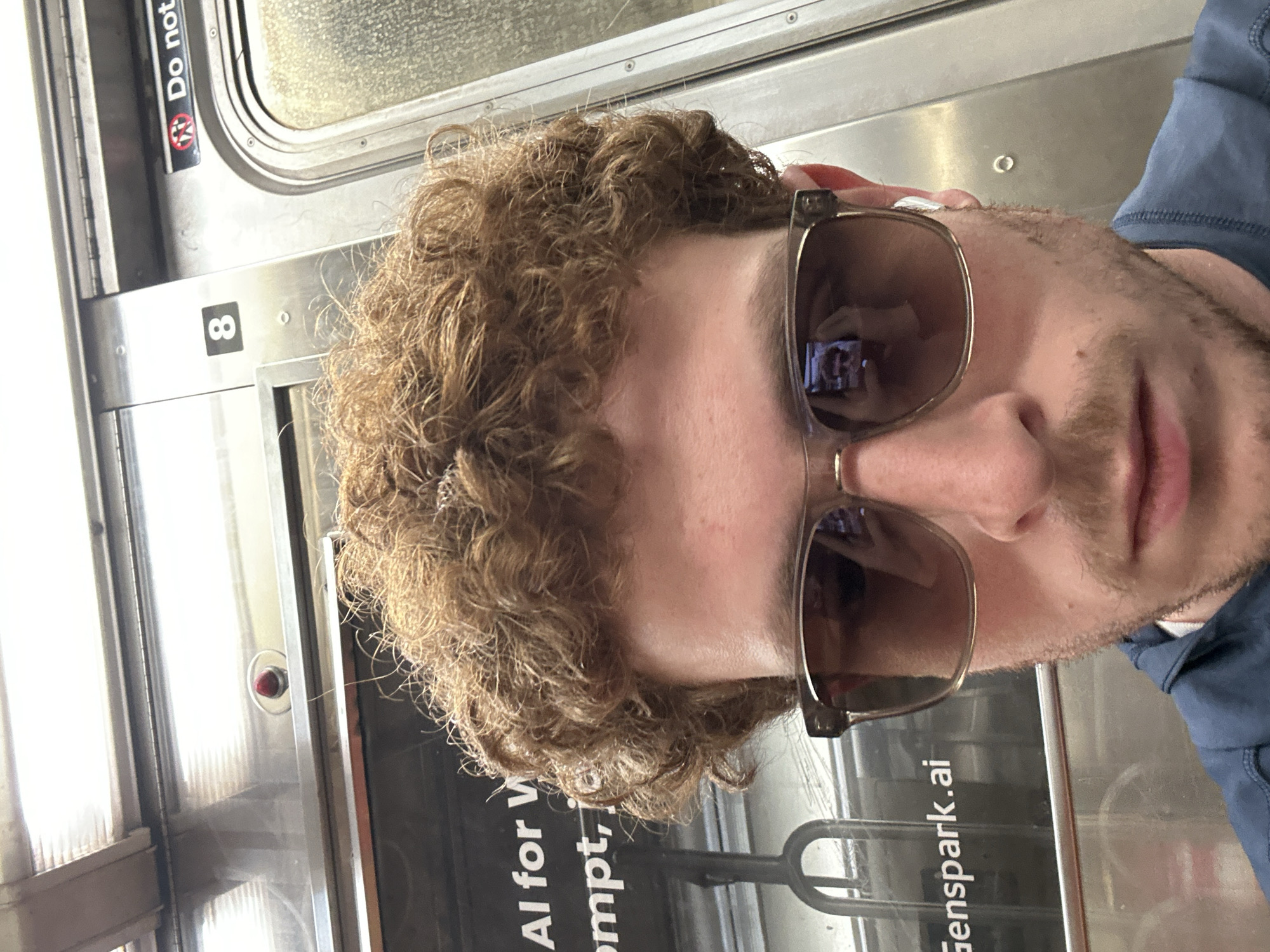 A close-up, portrait-style photograph shows a person with curly brown hair and light stubble, wearing sunglasses and a blue collared shirt. The person's eyes are obscured by the dark lenses of the rectangular, light-framed sunglasses. A reflection in the left lens reveals a bright screen, possibly a smartphone or monitor, displaying what appears to be text or an interface.
The setting is inside a public transportation vehicle, likely a subway or train car, in New York, United States of America. The background consists of metallic walls, a window with textured glass, and a door mechanism. A metal grab handle or pole is visible in front of the window.
Notable details on the subway car interior include a black-on-white sign with the number "8". Above it, another sign reads "Do not" followed by a symbol of a person with a diagonal line through it, likely indicating a restriction such as "Do not lean" or "Do not block." A small red light or button is also present near the door frame. Partially visible text on what might be an advertisement or display reads "AI for W", "ompt," and "Genspark.ai." The overall scene suggests the person is taking a selfie or being photographed while riding the subway. The time of day and specific weather conditions are not discernible, and the person's visible expression is neutral.