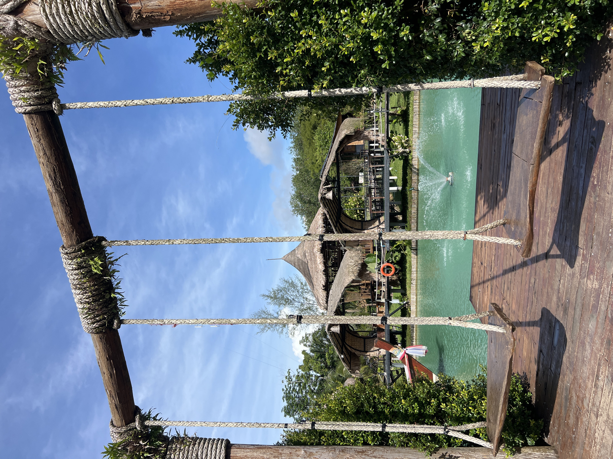 This image depicts a serene outdoor scene, likely a resort or restaurant, in Tambon Pa Kae Bo Hin, Thailand. In the foreground, two rustic wooden swings hang from a sturdy wooden beam supported by thick, rope-wrapped posts. The swings are suspended over a wooden deck. Beyond the deck, a vibrant turquoise body of water is visible, with a fountain spraying water in the center. In the background, several thatched-roof structures with open sides, typical of tropical architecture, are built over the water or on stilts. Lush green foliage and trees surround the area, and a clear blue sky with scattered clouds completes the picturesque setting. A small orange life preserver is hanging on a railing in front of one of the structures.