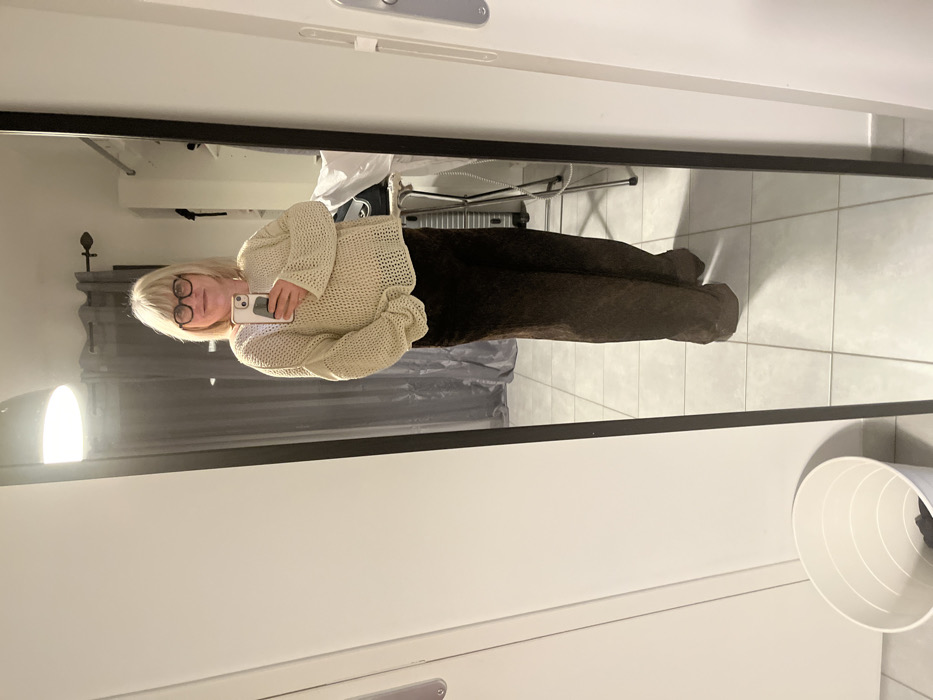 A mirrored image shows a person holding a phone, wearing glasses, a beige sweater, and dark pants. Behind the person are curtains and a suitcase. The mirror is framed with a black border. The surrounding environment includes white walls, a wastepaper basket, and tiled floors. The image is rotated 90 degrees.