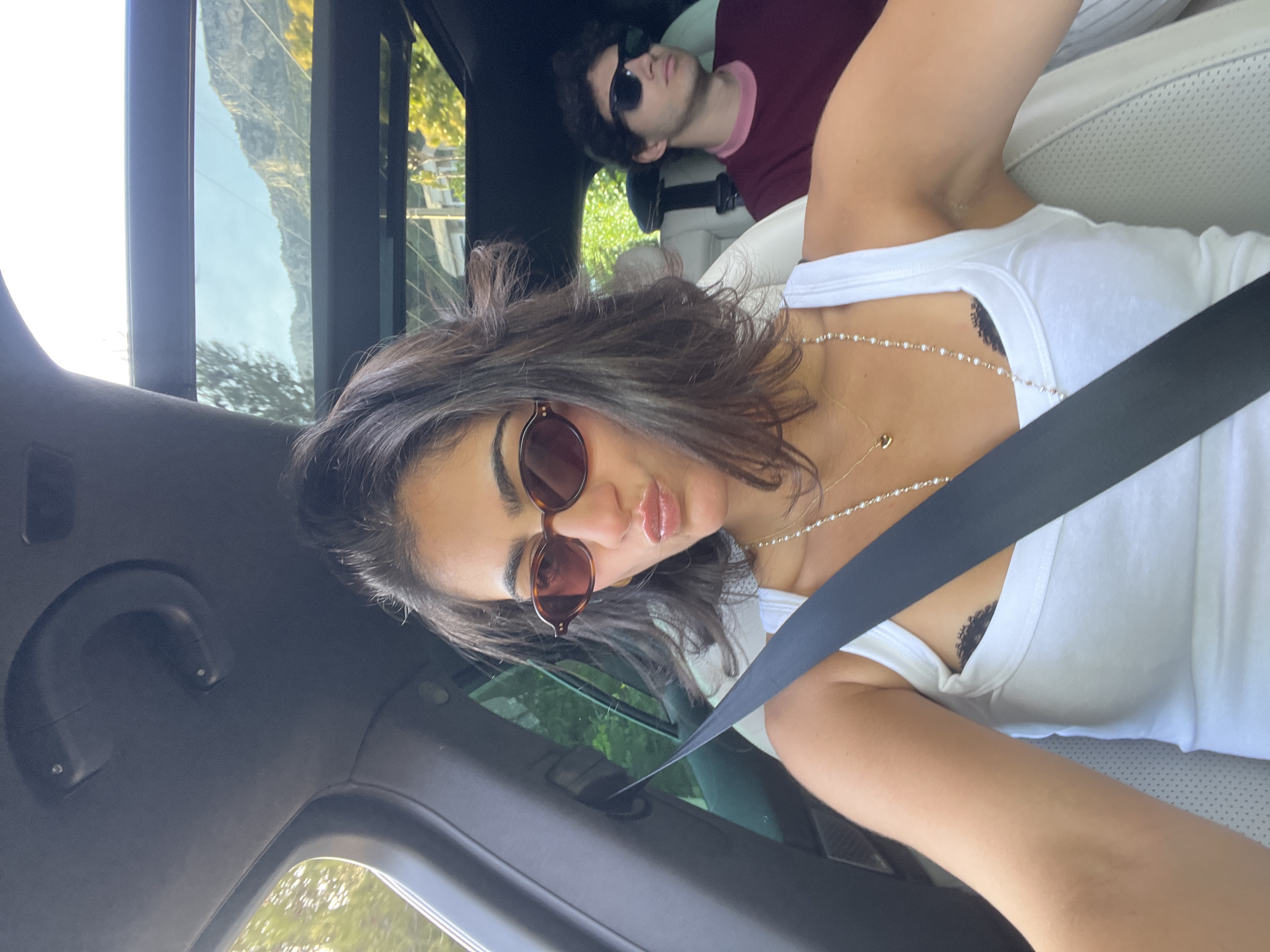 A woman wearing sunglasses and a white tank top, with a seatbelt across her chest, is taking a selfie inside a car. She is wearing a pearl necklace, and a tattoo is partially visible on her chest. Behind her and to the right, a man wearing sunglasses and a burgundy shirt is reclined in the passenger seat. The car's interior is predominantly black, with light-colored leather seats. Outside the car window, a mountain is visible, and greenery can be seen. The location is Èze, France.