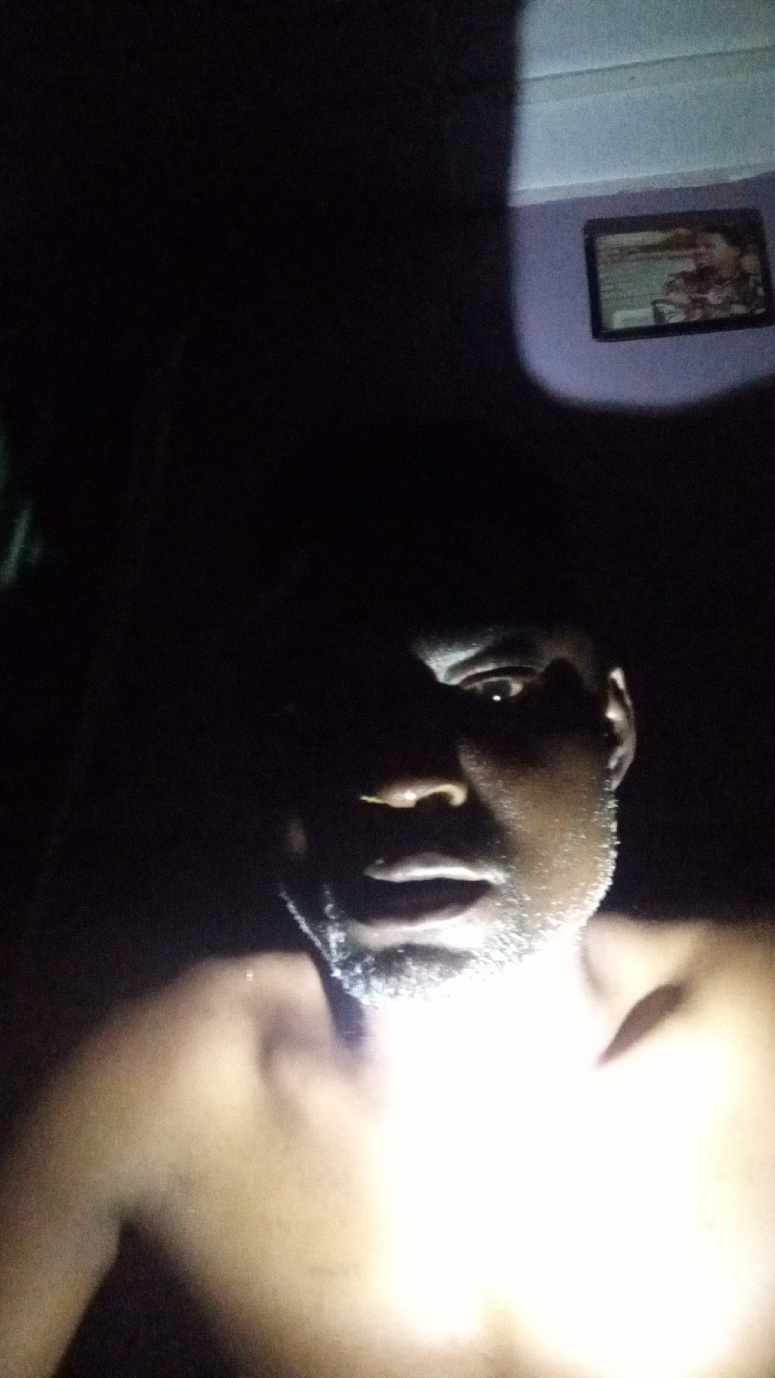A shirtless man with dark skin and a light beard is illuminated by a bright light from below. The background is very dark, but a purple wall with a framed picture can be seen in the upper right corner. The location, Sabon Tasha, Nigeria, is not discernible from the image.