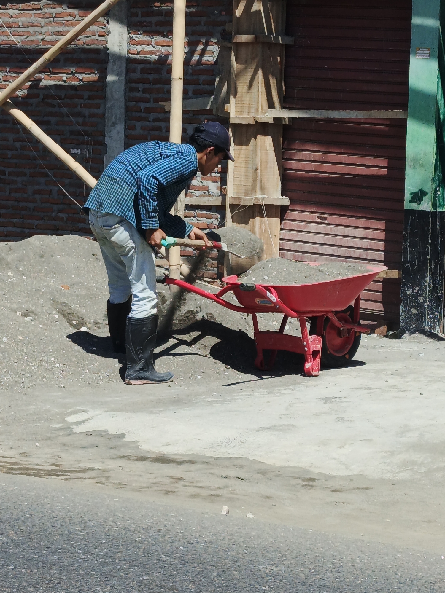 A man wearing a blue checkered shirt, light-colored pants, a black cap, and black rubber boots is using a shovel to load gravel into a red wheelbarrow. The wheelbarrow is filled with gravel. The man is standing on a dirt and gravel surface. In the background, there is a brick wall under construction with bamboo scaffolding. There is also a brown corrugated metal shutter and a green vertical beam. The scene appears to be a construction site in Kelurahan Cilacap, Kecamatan Cilacap, Indonesia.