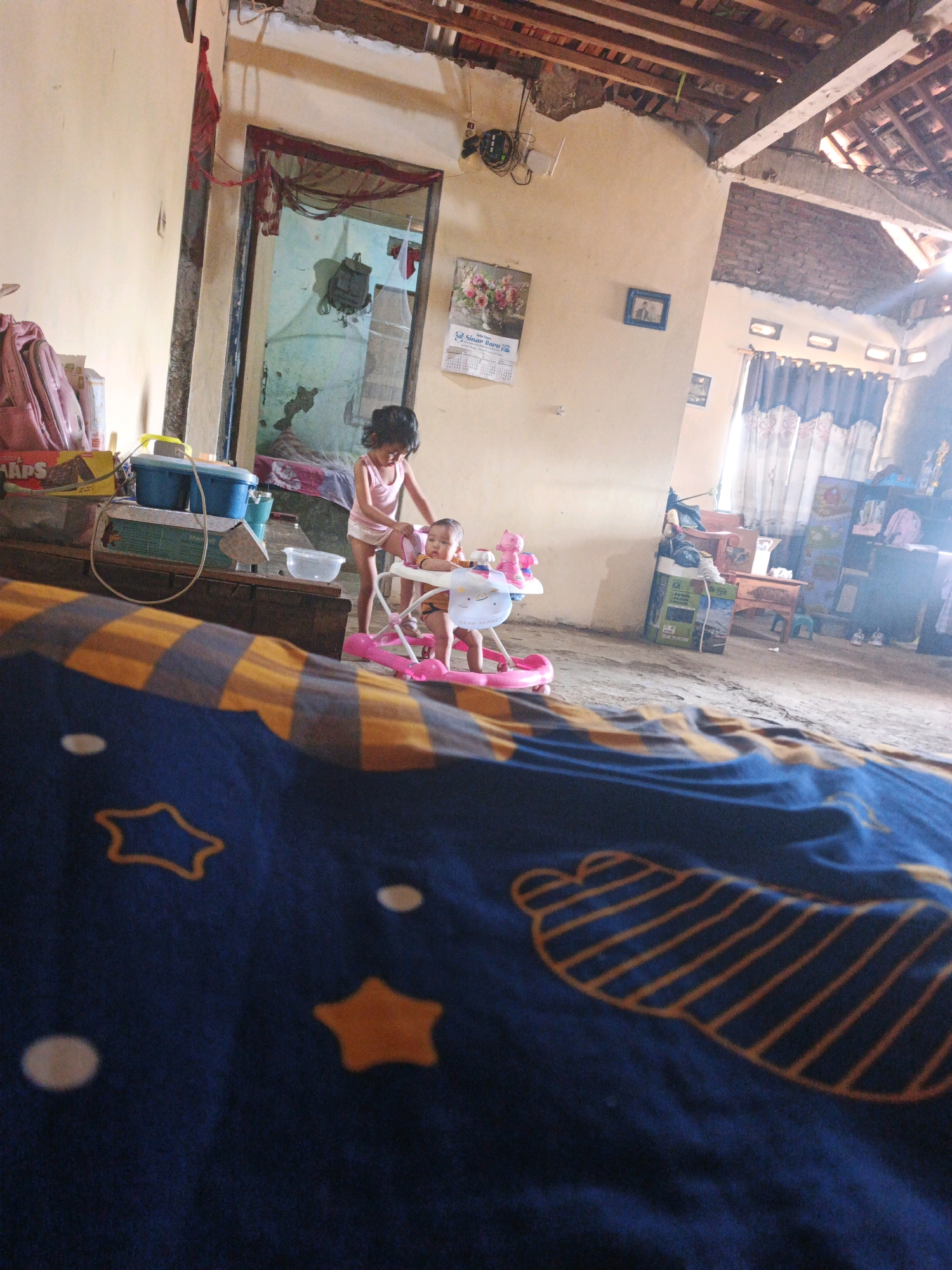 The image depicts an indoor scene, likely a home, with unfinished construction visible in the ceiling. In the foreground, a blue blanket with yellow and orange star patterns is draped across a surface. In the midground, two young children are interacting. A slightly older child, wearing a pink top and white bottoms, stands beside a pink baby walker. Inside the walker is a younger child, wearing an orange top and white bottoms. The environment appears modest, with concrete floors and light-colored walls. Various objects are present, including storage containers, a backpack, and a calendar on the wall. The overall impression is of a lived-in, somewhat unkempt domestic space. The scene is consistent with a residential setting in Bumiharjo, Indonesia.