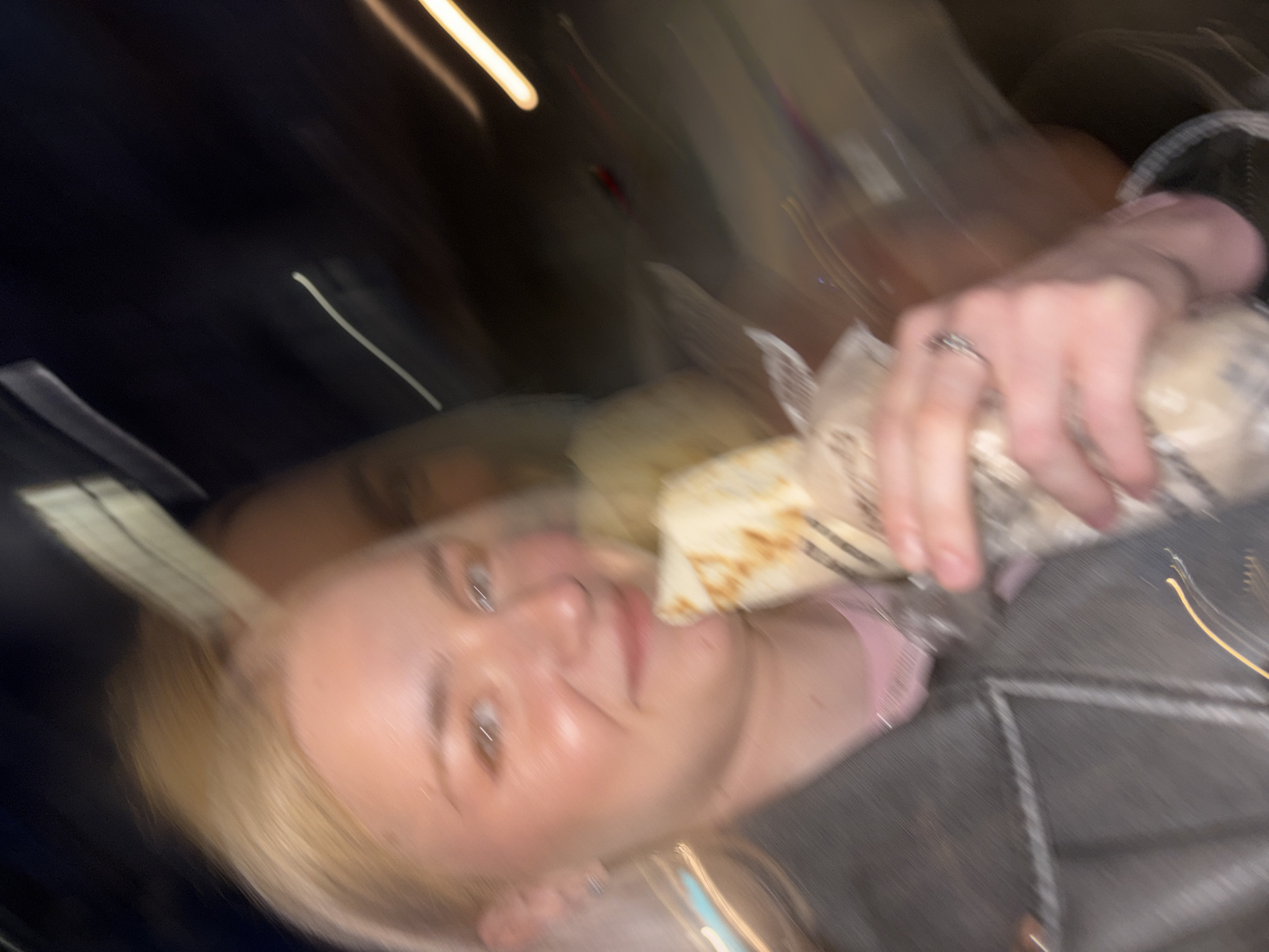 The image shows a blurred frontal shot of a young Caucasian woman with blonde hair, holding a wrapped food item, possibly a burrito or kebab, in her right hand. She is wearing a dark jacket over a pink shirt, and a ring is visible on her right hand. The background is dark and blurred, with streaks of light suggesting movement or a night setting, potentially indicating a street scene in Saint Petersburg, Russia, with its characteristic urban lighting and dark ambiance. The overall impression is that of a candid, on-the-go snapshot.