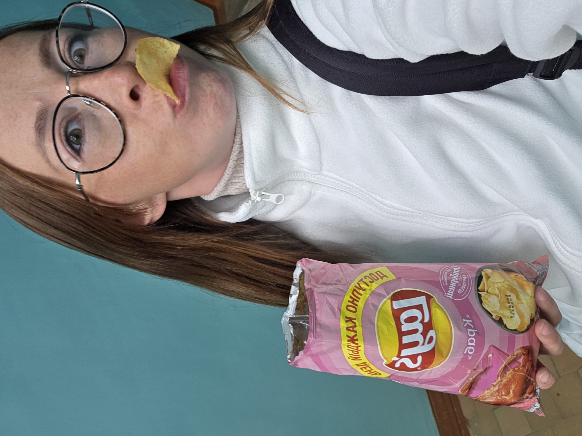 A rotated medium shot shows a Caucasian woman with brown hair and wearing round glasses. She has a single potato chip stuck to her upper lip. She wears a white fleece jacket over a turtleneck and a black backpack. She holds a bag of Lay's "Crab" flavored potato chips, printed in Cyrillic. The background is a pale turquoise wall and part of a wooden doorway is visible. The location is Chaikovsky, Russia, given the Cyrillic on the Lay's bag.
