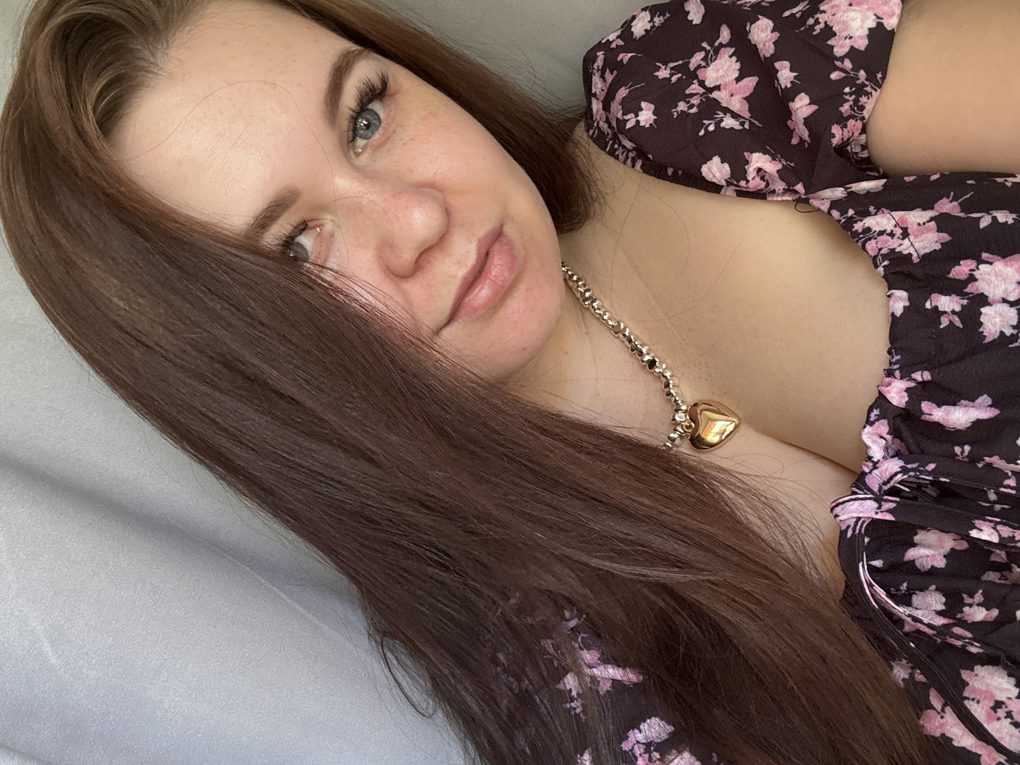 The image shows a close-up, angled selfie of a young woman with long, dark brown hair and blue eyes. She has a light complexion with freckles visible on her cheeks and nose. Her hair is dark brown and falls over her face, partially obscuring her left eye. She is wearing a dark floral print dress with pink flowers and a gold heart-shaped pendant necklace. The background appears to be a light-colored, possibly fabric surface, suggesting an indoor setting. The lighting appears soft, potentially indicating natural light. Her expression is relaxed and slightly sensual. No text is visible in the image. The location context is Chaikovsky, Russia, but there are no specific visual cues in the image to confirm this.