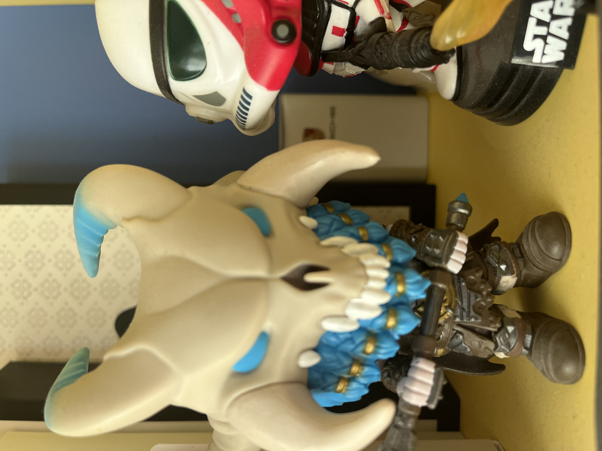 An indoor close-up shot captures two collectible figures displayed against a mixed background. The image is oriented vertically, with the figures appearing sideways or inverted relative to each other.
In the upper portion of the frame, an inverted Star Wars clone trooper figure is visible. It features a white helmet with a dark green visor, and its armor is detailed with red and black accents. Part of its black base, which clearly displays the white "STAR WARS" logo, is also visible. The clone trooper appears to be hovering or mounted above the second figure.
The main subject of the image is a larger, fantastical figure positioned in the foreground and lower half of the frame. This figure has a light, bone-colored head with prominent, curving horns/tusks that have light blue tips and ridges. Its stylized face features small blue eyes. Around its neck or shoulders, there's an elaborate blue and white decorative collar or mane, adorned with golden bands or details. The figure's body is depicted in shades of brown and grey, featuring intricate armored textures and details, and it appears to be holding a dark staff or weapon with light-colored hands.
The figures are arranged on a flat, light yellow surface, possibly a shelf or display stand. The background behind them is a solid blue wall or surface, with a section on the left showing a light-colored surface with a repeating geometric pattern, suggesting wallpaper or a patterned box. The lighting is consistent, indicating an indoor setting.