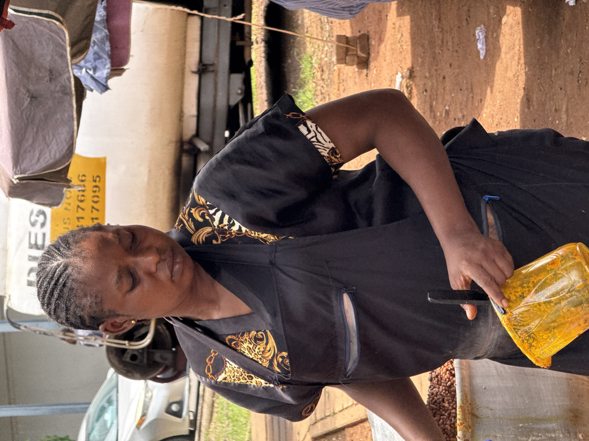 In Abuja, Nigeria, a woman wearing a black apron with gold trim stands outdoors near a food stall. The apron has a zippered pocket. She is holding a yellow plastic cup filled with an orange-colored substance, possibly food or a sauce. In the background, a large white truck with yellow signage and a silver car are visible. The ground is covered in reddish-brown dirt. The lighting suggests it is daytime.