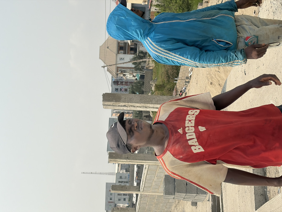This image captures an outdoor scene in Alaguntan, Nigeria, on what appears to be a construction site or undeveloped land under a bright, hazy sky.
Two adult males are present. In the foreground on the left, an individual with a dark complexion wears a black baseball cap with a white Nike swoosh and a soiled red and off-white raglan-sleeve t-shirt featuring an Adidas logo and "BADGERS" with a stylized 'W' in white text on the chest. His face is visible, displaying a slight grimace or squint, and he is looking towards the right. His right arm is partially visible, extended downwards.
To the right and slightly behind the first individual, a second male is partially visible. He is wearing a blue hooded jacket with three white stripes on the sleeves and light-colored, soiled pants. His face is obscured by the raised hood. A yellow and black retractable tape measure is clipped to his right hip. His right hand is visible, appearing to hold a small object.
The immediate environment consists of sandy/dirt ground. To the left, a partially constructed wall made of concrete blocks is visible, alongside a stack of new concrete blocks. Several raw concrete pillars stand vertically in the foreground and mid-ground. In the background, various buildings are present, including multi-story structures and a prominent two-story house with a light brown gabled roof. A communication tower or similar structure is visible in the far distance on the left horizon. Scattered refuse and sparse vegetation are also visible in the mid-ground.