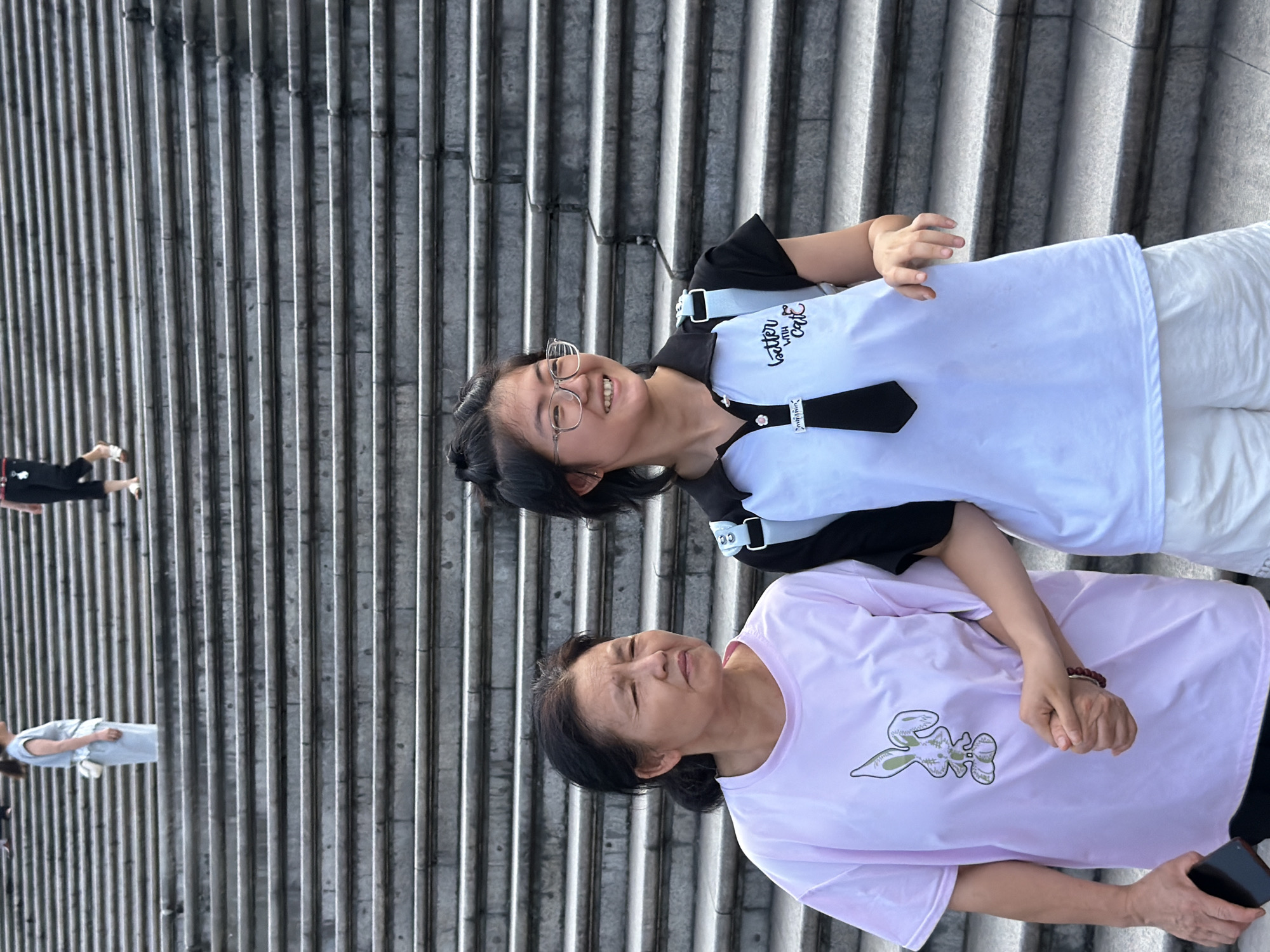 Two women are standing on a wide outdoor staircase in Yuzhong District, China. The woman on the left, appearing to be older, wears a light pink t-shirt with a graphic of a rabbit. She is holding hands with the younger woman to her right. The younger woman, wearing glasses, is dressed in a white polo shirt with black accents and a black tie, and white shorts. She is looking to her right and smiling. The staircase is made of concrete and appears to be quite long. In the background, further up the stairs, other individuals can be seen, including one in a light blue outfit and another in black. The lighting suggests it is daytime.