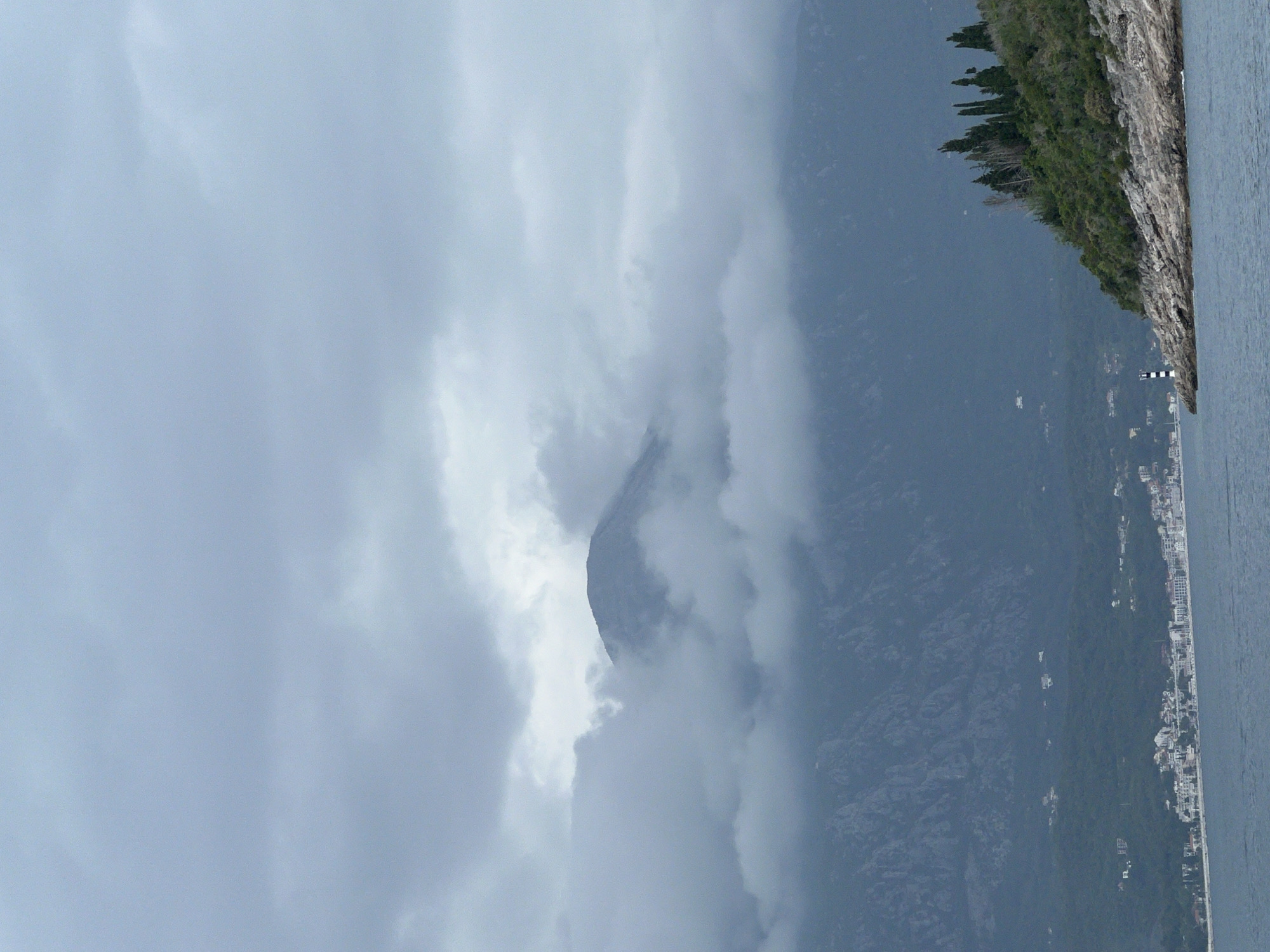 The image captures a cloudy day on the coast of Luštica, Montenegro. A craggy mountain peak is partially obscured by heavy clouds. To the right of the mountain, the coastline is lined with dense green vegetation atop rocky cliffs. Further along the coast, a string of buildings suggests a town or resort area. In the distance, a narrow body of water is visible.