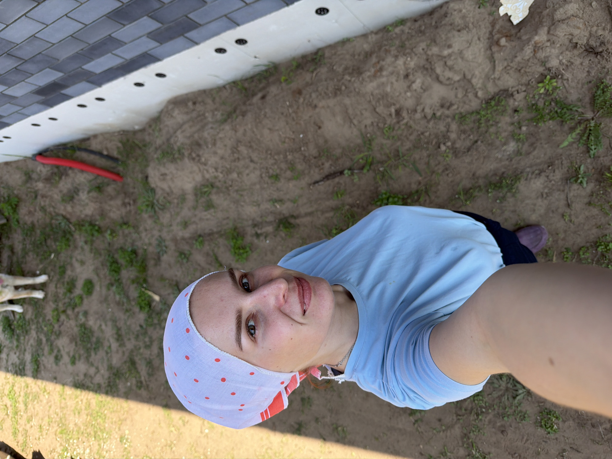 A woman in a light blue t-shirt and a white headscarf with red polka dots is taking a selfie outdoors. She is standing on dirt ground with sparse grass. In the background, there is a building foundation with grey brick paving on top and a white base. A red and black cable runs along the base. The scene is located in Chaikovsky, Russia. A partial view of a white and brown dog can be seen on the left side of the frame.