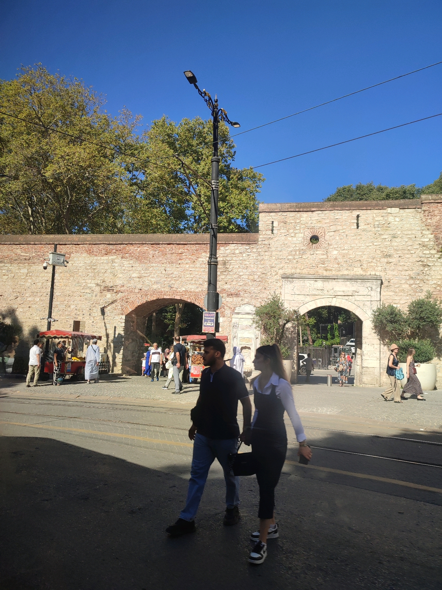The image depicts an outdoor scene in Istanbul, Turkey, likely in the vicinity of historical walls and arches. In the foreground, a man and a woman walk on a paved street, their figures casting long shadows. The man is wearing a dark t-shirt and blue jeans, while the woman is in a white collared shirt and a dark pinafore dress. Behind them, a cobblestone path leads to an arched gateway in a substantial stone wall. Several people are visible near food stalls and in the background, with one stall selling what appears to be simit, a Turkish bread. Trees with green foliage are present above the wall, and a clear blue sky dominates the upper portion of the frame. A lamppost stands near the foreground, and power lines are strung across the sky. The architecture suggests a historical setting, possibly within the city walls.
