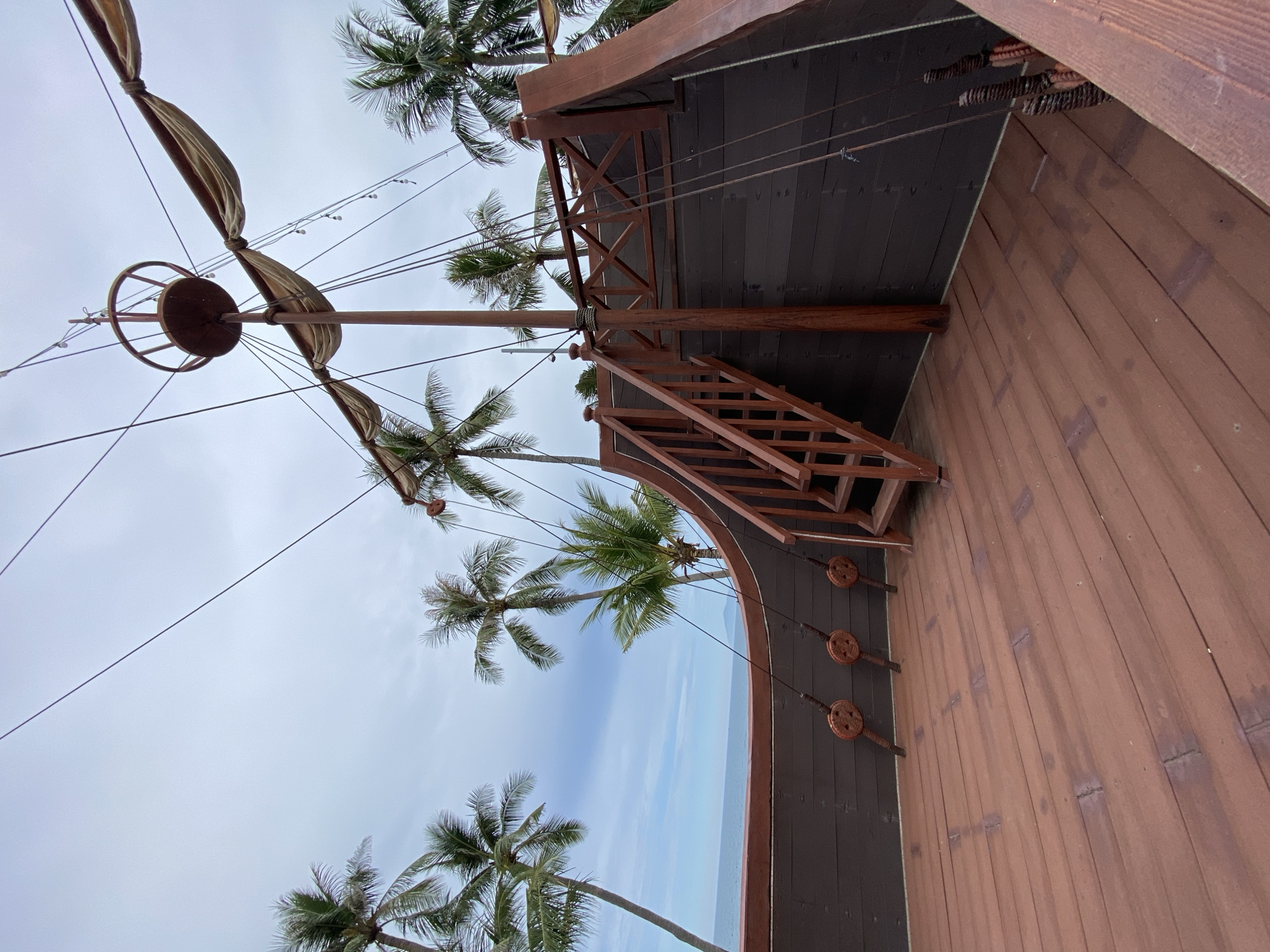 The image depicts a section of a wooden pirate ship replica, situated outdoors with palm trees and the ocean in the background. The ship's deck is made of weathered brown planks, and a wooden staircase leads up to an upper level. The ship features a prominent mast with rigging and a crow's nest, along with parts of sails. Decorative rope-wrapped spools are visible along the ship's hull. The scene is set against a cloudy sky, with the ocean visible beyond the ship's railing. The presence of palm trees suggests a tropical coastal location. This pirate ship replica is located at VinWonders Nha Trang, an amusement park and resort complex on Hon Tre Island, off the coast of Nha Trang, Vietnam.