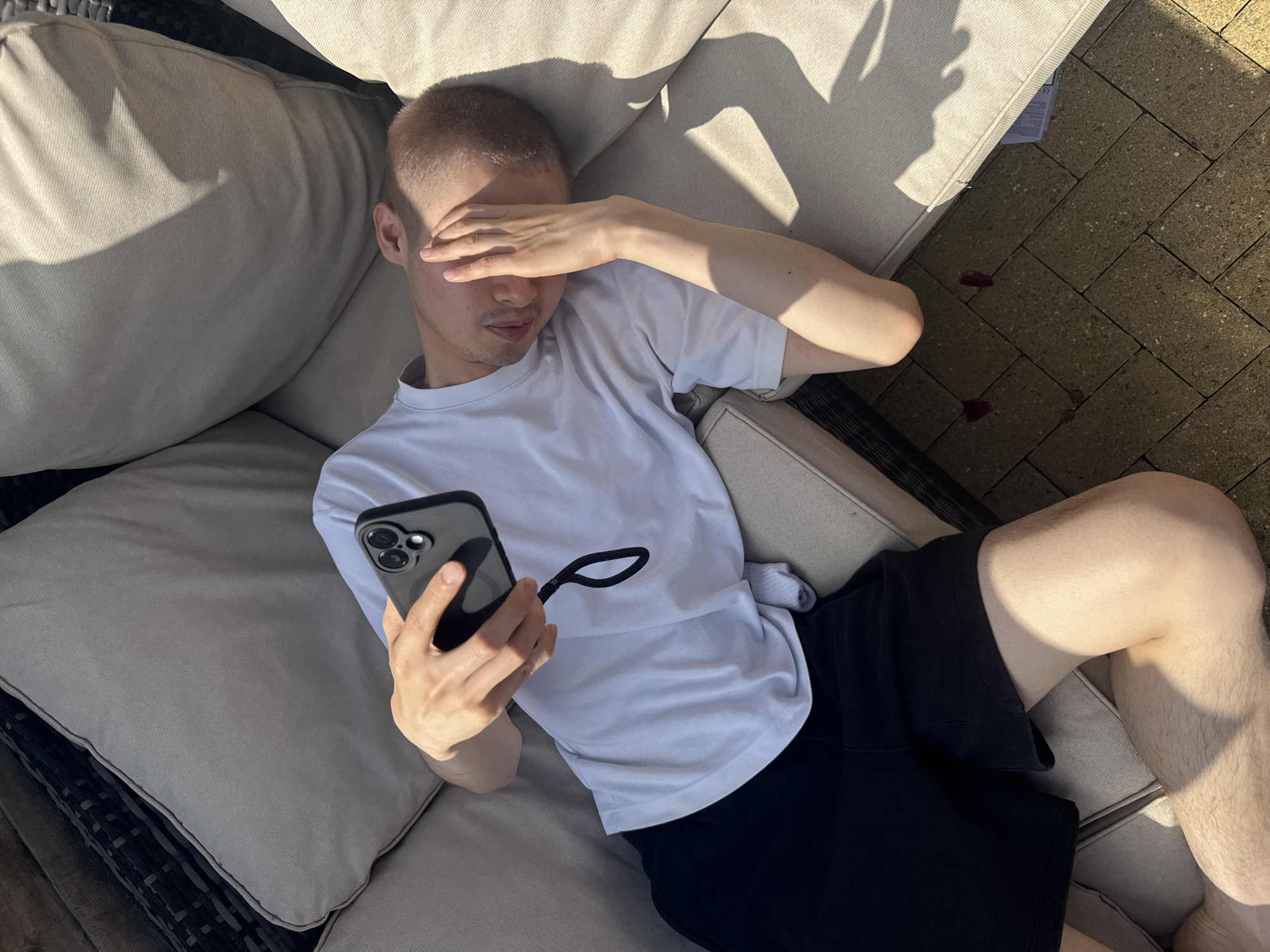 The image shows a person lying on a couch outdoors, with their hand covering their eyes and holding a smartphone. The person is wearing a white t-shirt and black shorts. The couch appears to be part of an outdoor furniture set, possibly on a patio or in a garden. The ground is paved with rectangular stones. Sunlight casts shadows on the couch and ground. There are a few red petal-like objects on the ground near the couch. The scene is set in a residential area, likely a garden or patio, which is commonly found in cities like London, United Kingdom. There is no specific indication of the precise location within London, but the overall setting suggests a domestic outdoor space.