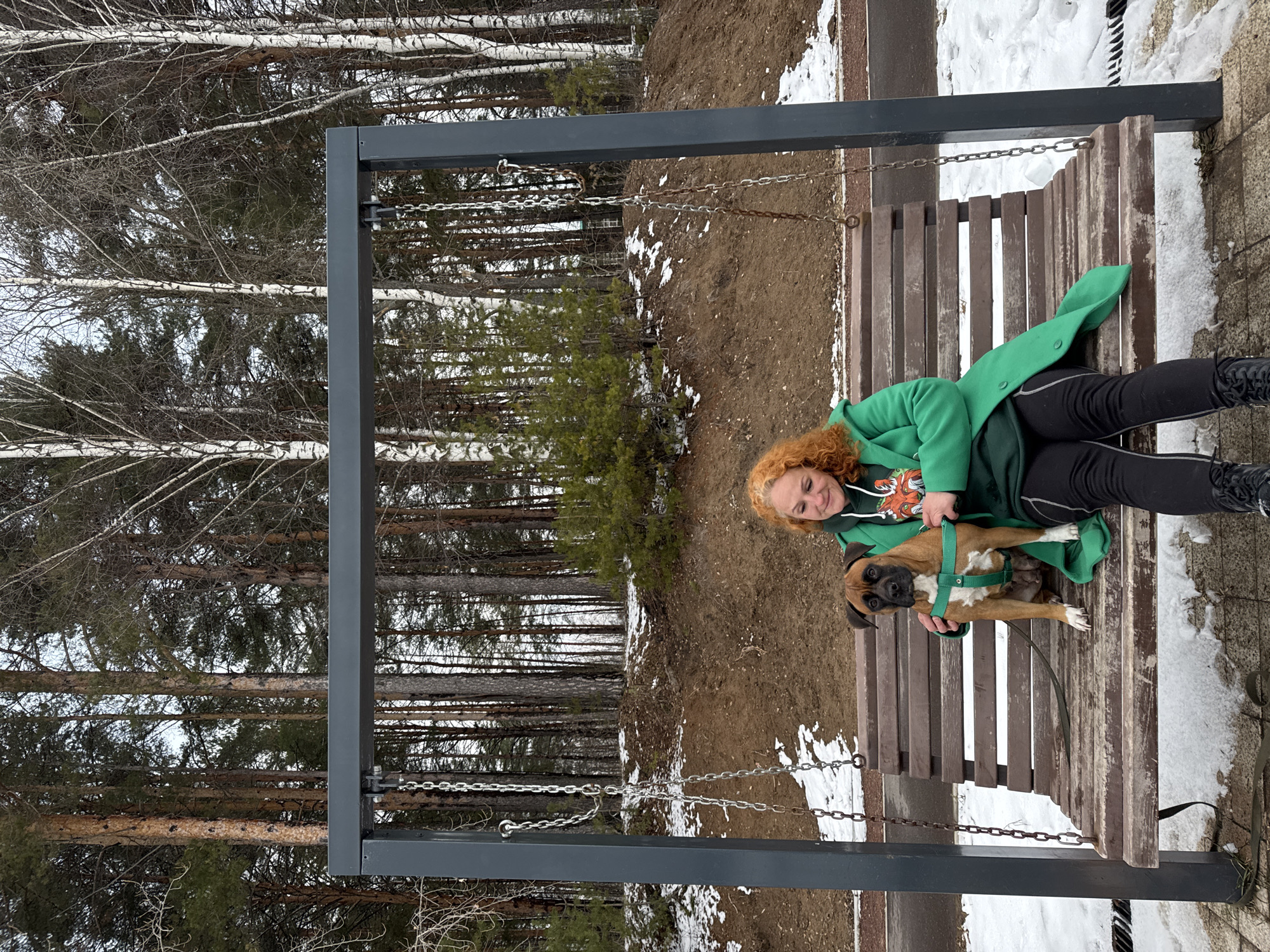 A woman with curly red hair and wearing a green coat sits on a wooden swing with a light brown boxer dog with a green harness in her lap. The swing is suspended by metal chains from a dark gray frame. The background includes a forest of trees, some with white bark, and patches of snow on the ground. A paved area is visible to the side of the swing, also with some snow. The photo was taken in the city of Chaikovsky, Russia.