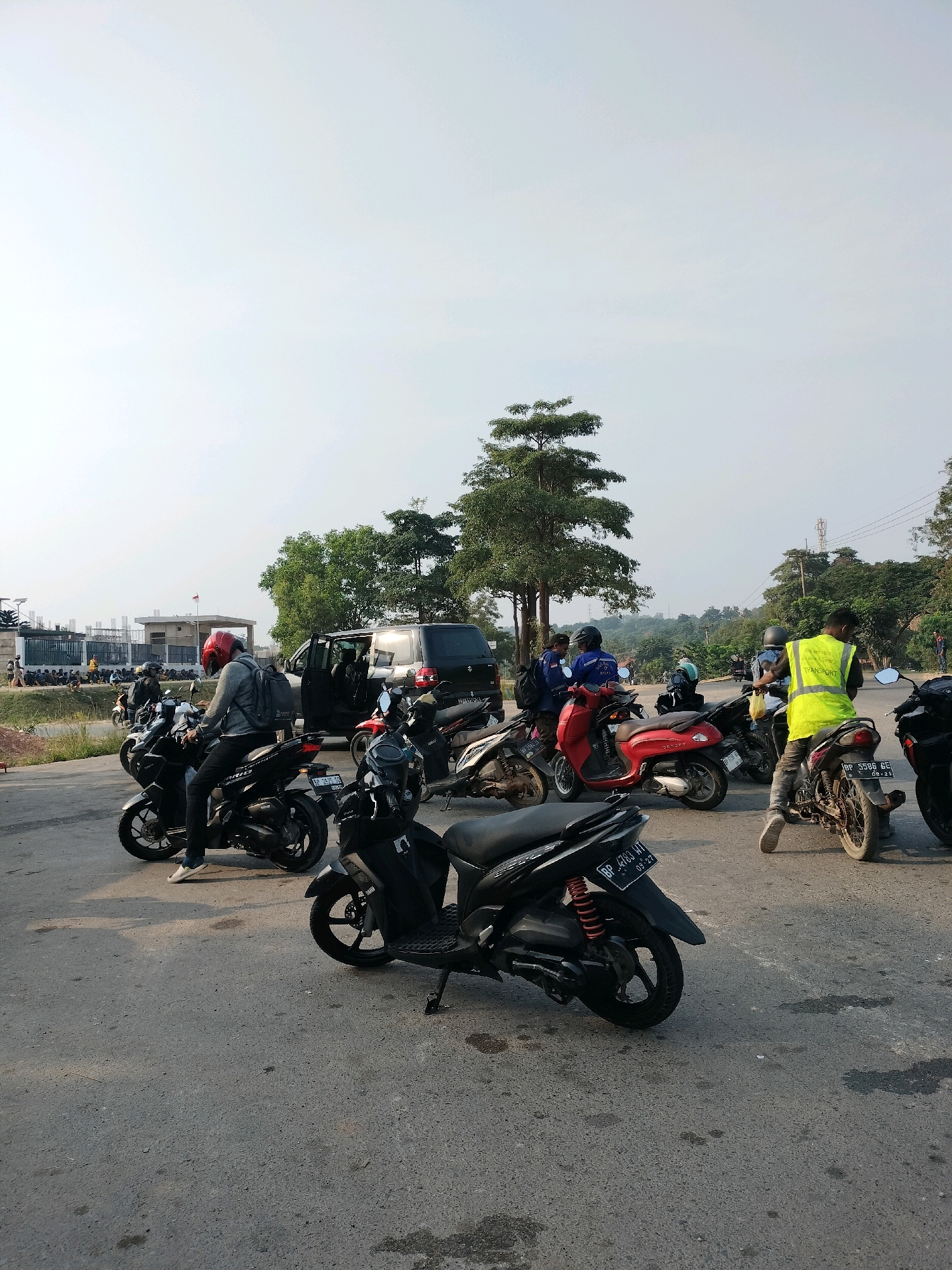 The image depicts a scene in Batam, Indonesia, likely an industrial or construction area, given the presence of scaffolding and unfinished buildings in the background. The foreground is dominated by a gathering of motorcycles and several individuals, suggesting a temporary stop or assembly point.
In the center of the frame, a person wearing a red helmet and a grey jacket sits on a black motorcycle. Another individual in a bright yellow vest, with "All Service Staff Client Transport" printed on it, is seen from behind, also near a motorcycle. Several other motorcycles are parked or being operated by individuals dressed casually. A black van with its doors open is parked to the left, with people around it.
The environment appears to be an asphalt surface, possibly a road or parking lot, with some scattered debris and oil stains. The background features trees and the hazy sky typical of tropical climates, with construction elements visible in the distance. The overall impression is one of an informal gathering of workers or commuters in an urban or peri-urban setting.