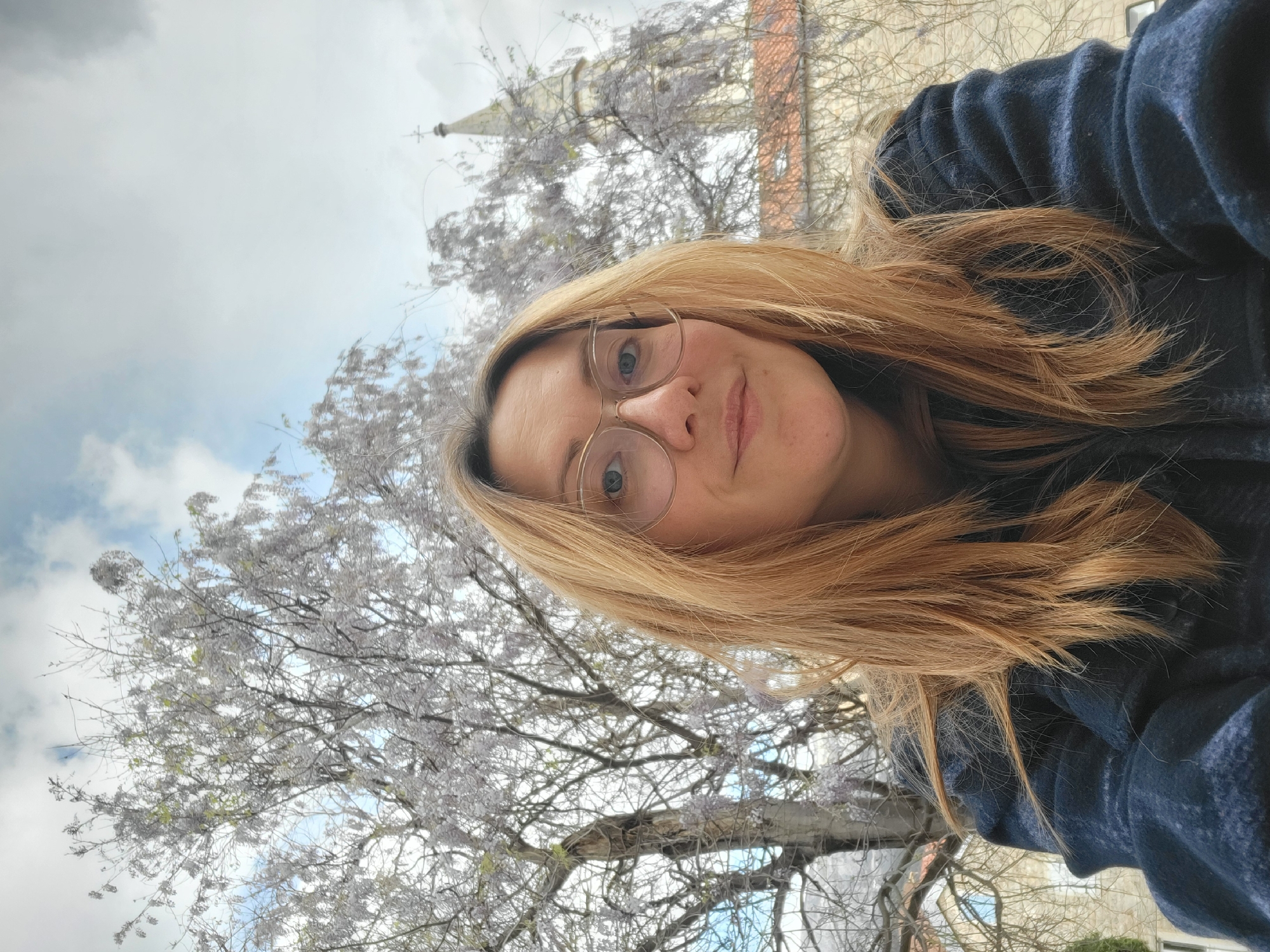 A woman with glasses is taking a selfie in Budva, Montenegro. Behind her is a tree with white flowers, and a tall building in the background. The sky is partly cloudy. She is wearing a dark blue sweater.