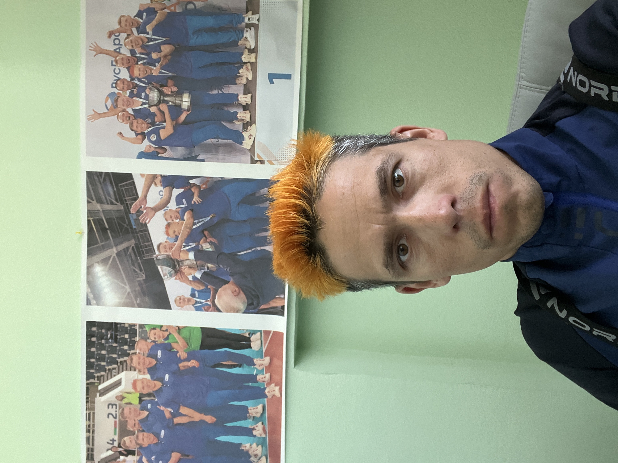 A medium shot captures a man with orange-dyed hair. He is positioned to the right of the frame, wearing a dark blue top with a contrasting dark trim bearing the word "NORTH". Behind him, affixed to a light green wall, are several photographs of a sports team wearing blue uniforms. A white-colored seat is visible in the top right corner. Based on the information present on the posters, the location is likely the city of Chaikovsky, Russia.