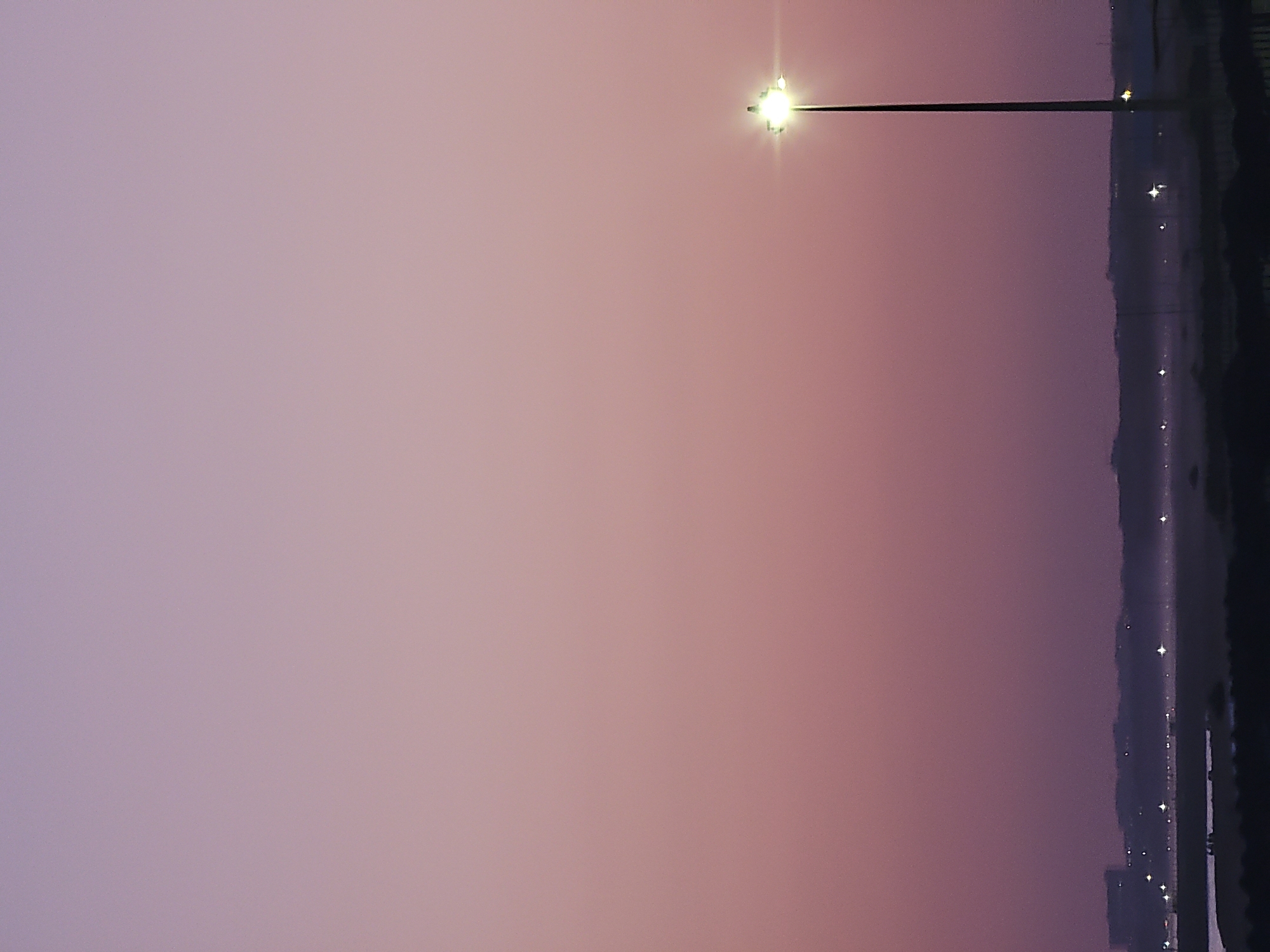 The image depicts a twilight scene in Quarteira, Portugal, characterized by a soft, gradient sky transitioning from a pale lavender at the top to a dusky purple near the horizon. A solitary tall lamppost stands prominently on the right side of the frame, its bright light illuminating the surroundings and creating a starburst effect. In the distance, a hazy, silhouetted shoreline is visible, dotted with faint lights that suggest buildings or other urban features. The foreground is dominated by dark, indistinct landmasses, possibly marshland or a beach, with a sliver of water reflecting the subdued sky. No people are visible in the image. The overall atmosphere is serene and quiet, emphasizing the fading light of day and the nascent glow of artificial illumination.