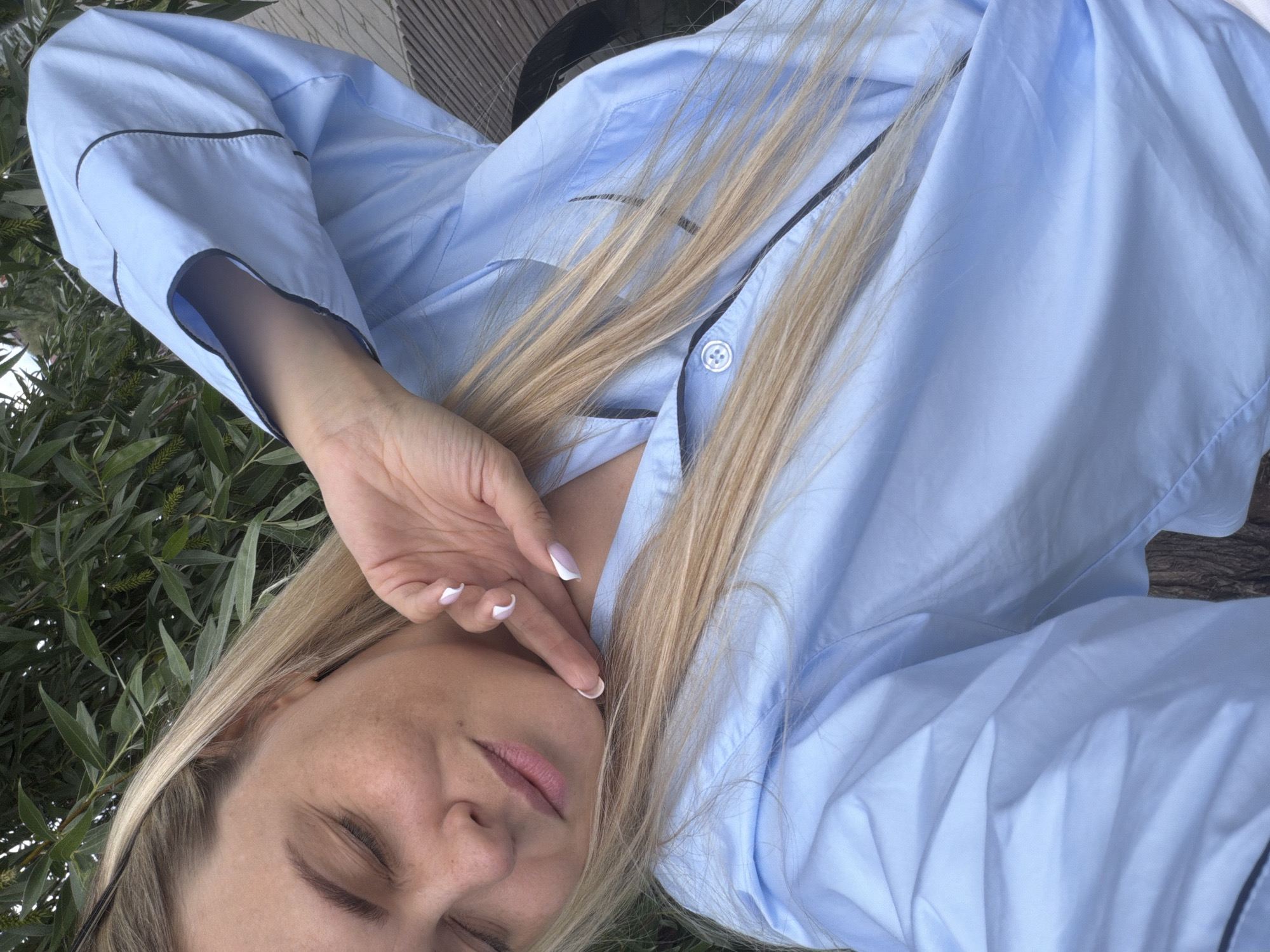 This image captures a close-up, overhead view of a woman with long, straight blonde hair, resting outdoors in what appears to be Votkinsk, Russia. She is wearing a light blue button-up shirt, possibly a pajama top, detailed with dark piping along the edges and a visible white button on the placket. Her eyes are closed, conveying a sense of peace and relaxation. One hand, adorned with a white-tipped French manicure, is gently placed near her face, with a finger touching her cheek.
The setting is an outdoor environment, evident from the soft, natural daylight and the vibrant green foliage, possibly willow leaves, that frames the left side of the frame and appears in the background. A textured wooden surface, which could be part of a deck or outdoor furniture, is visible in the upper background. The lighting suggests it is daytime, with no indications of specific weather conditions other than clear skies allowing for bright illumination. The overall mood of the image is calm and tranquil, highlighting a moment of rest or contemplation. There is no visible text in the image.