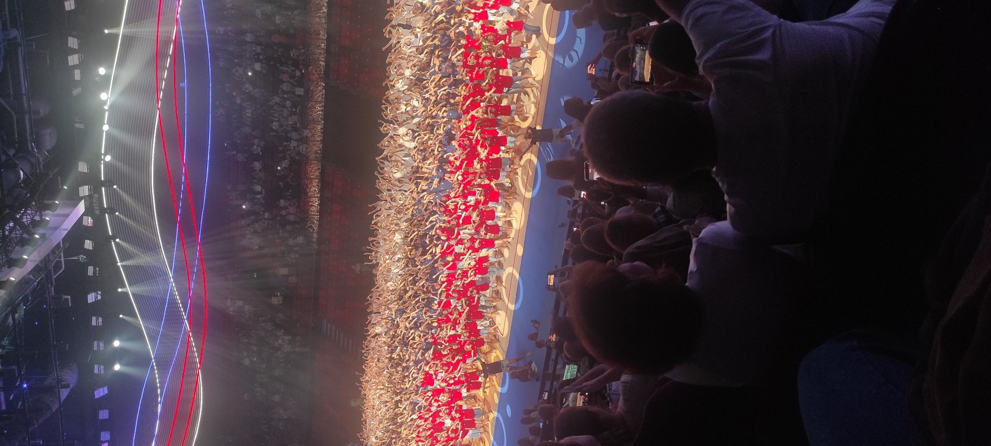 The image captures a large-scale performance event, likely a concert or theatrical production, taking place indoors within a stadium or arena. The scene is divided into two main areas: the performance stage and the audience seating.
On the stage, a vast crowd of individuals, predominantly young people, are organized in rows. They are wearing matching red t-shirts and shorts, suggesting they are part of a unified performance group. Many of them are performing synchronized movements, with their arms raised in unison. The stage itself is brightly lit, with beams of light illuminating the performers from above. Behind the performers, there is a backdrop with a visually striking design of undulating red, white, and blue lines, possibly illuminated LEDs or projections, creating a dynamic visual effect. The floor of the stage is dark, with circular light patterns visible in the foreground, likely part of the stage's lighting design.
In the foreground and extending into the midground, the audience is seated in tiered seating that wraps around the performance area. The audience members are largely in silhouette due to the lighting directed at the stage. Many are holding up mobile phones, presumably to record the performance. The seating appears to be a dark color, possibly upholstered or made of plastic. The overall atmosphere suggests a well-attended event with a focus on a vibrant, energetic performance.
The location is identified as Moscow, Russia. While the specific venue is not explicitly stated, the scale and design suggest a major indoor arena like the VTB Arena or Luzhniki Palace of Sports, which are known for hosting large concerts and events in Moscow. The lighting and stage design are consistent with modern performance venues.