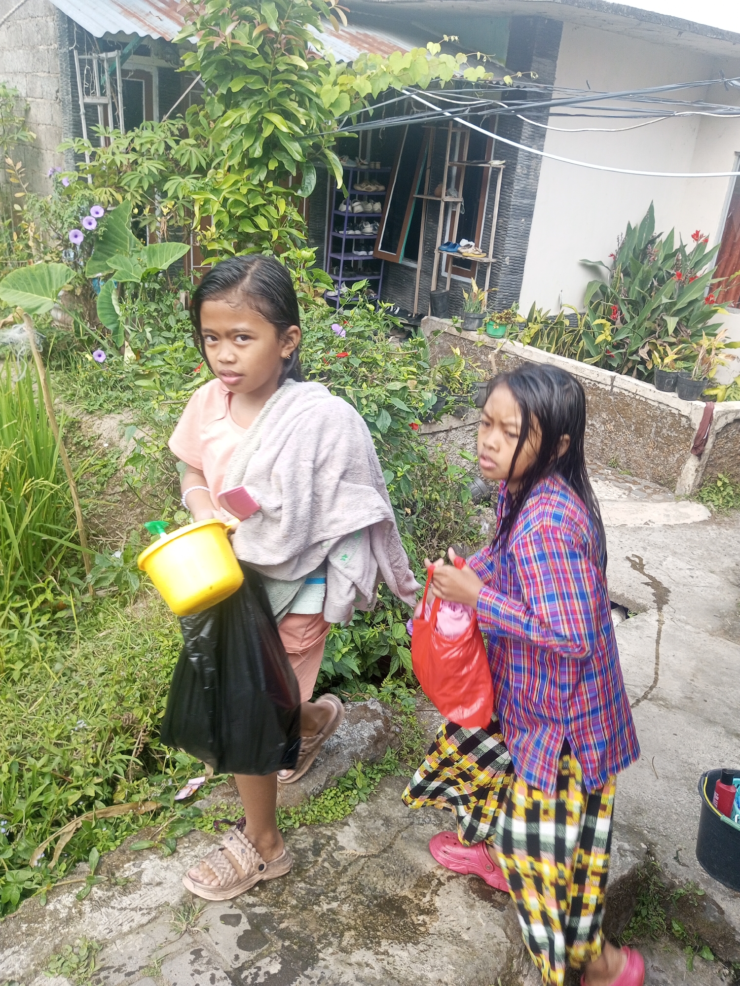 Two young girls are walking on a wet, paved pathway in front of a building that appears to be a residential home. The building has a corrugated metal roof and a light-colored facade with some stone accents. Green foliage and plants surround the area, including some flowering plants with purple and red blooms.
The girl on the left is wearing a light orange t-shirt and a towel draped over her shoulders. She is carrying a yellow bucket with a black plastic bag hanging from it. She is also wearing light brown sandals.
The girl on the right is wearing a colorful plaid long-sleeved shirt and a matching plaid skirt. She is carrying a red plastic bag in her right hand and is wearing pink slip-on shoes.
The ground is wet, suggesting it may have recently rained or been watered. In the background, there is a shoe rack with several pairs of shoes. A black bucket with some items inside is visible to the right of the second girl. The overall scene suggests an outdoor setting in a residential area, possibly in a rural or suburban part of Krasak, Indonesia, given the presence of lush vegetation and the style of the building.
