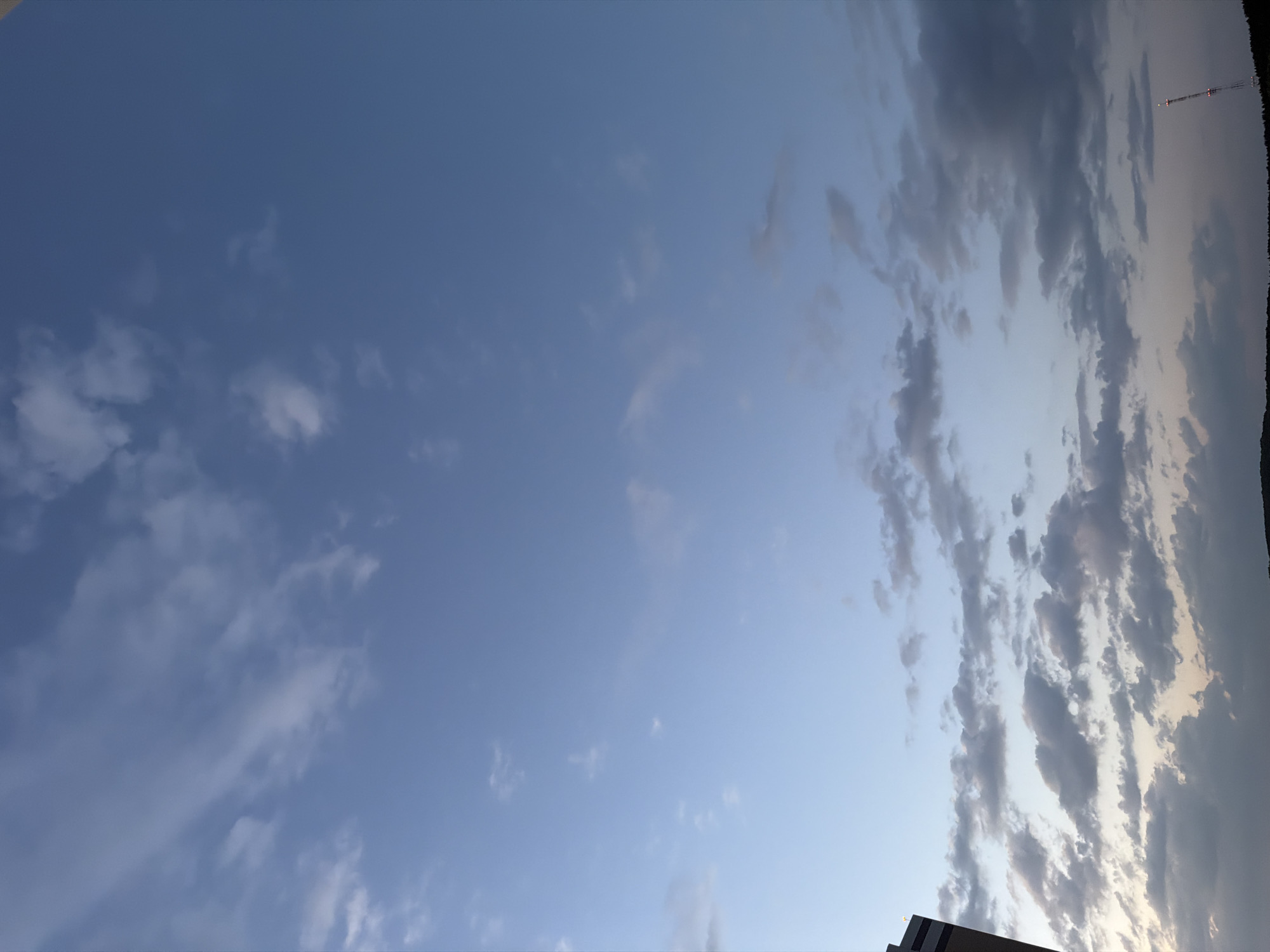 The image captures a view of the sky, which transitions from a clear, light blue on the left side of the frame to a more overcast area on the right. The right side features a dense array of striated clouds, ranging in color from light gray to darker shades, suggesting varying levels of sunlight and shadow. In the top right corner, a tall communications tower is barely visible against the sky. In the bottom right corner, a small part of a building is visible.