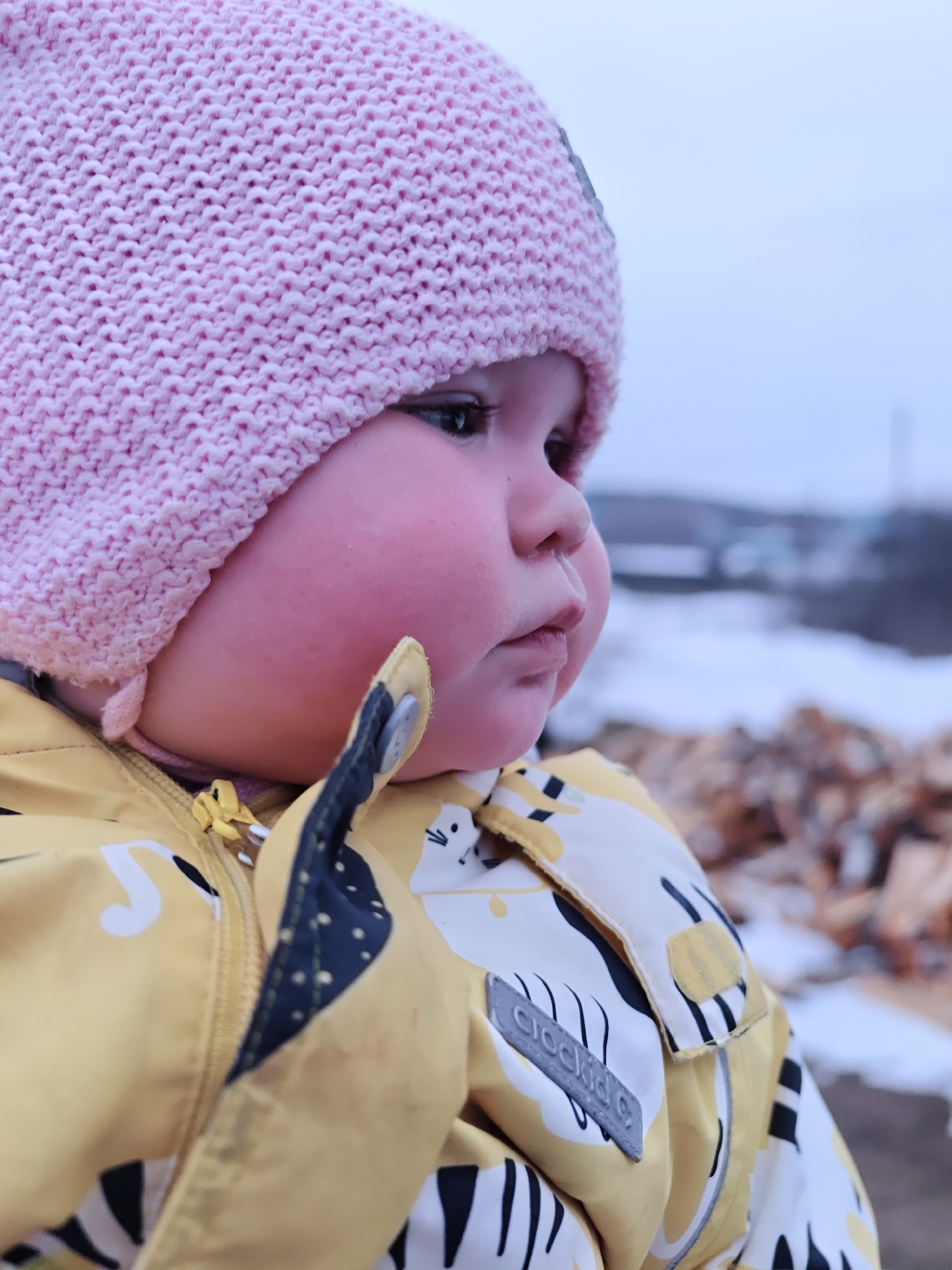 In a medium shot, a young child wearing a light pink knit hat and a yellow patterned jacket is seen in Уральское, Russia. The child's face, especially the cheeks, appears rosy. The jacket features a pattern of white and black on a yellow background, with a "Crockid" brand label visible. The background is slightly blurred, showing a landscape with patches of snow and a pile of wooden logs. Buildings and trees are visible in the distance under a cloudy sky.