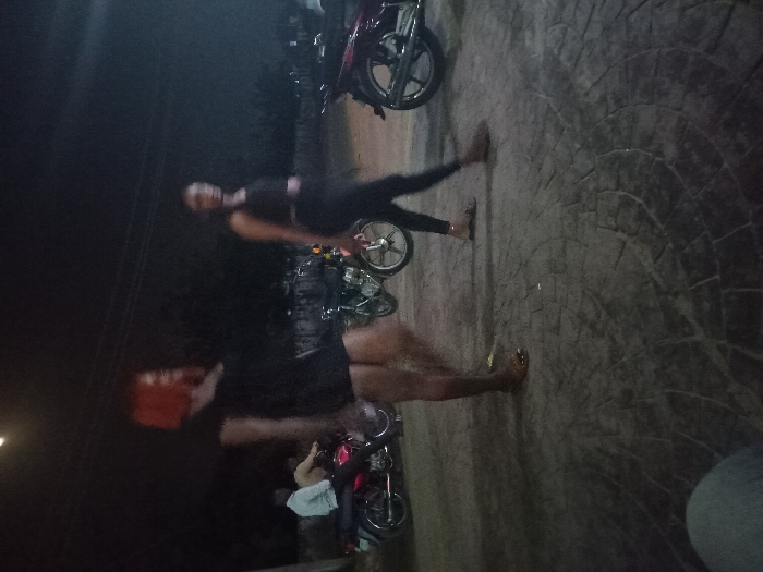 Two individuals appear, captured on a dark, paved surface alongside parked motorcycles. The figure on the upper side is wearing black clothing and seems to be in motion, while the other individual on the bottom side is partially obscured but also dressed in black. The background is dark, with the paved surface showing a circular pattern design.