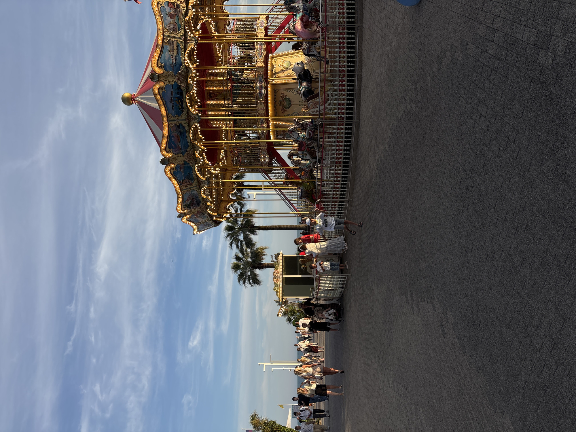 A carousel is operating on a paved walkway in a city park in Sochi, Russia, under a partly cloudy sky. People are walking along the walkway, with some gathered near the carousel. The carousel is lit with many bulbs and features ornate paintings on its canopy. Palm trees are visible behind the carousel and people. A fence surrounds the carousel, and a small ticket booth is situated nearby. The ground is paved with interlocking bricks. The overall scene suggests a leisurely afternoon or evening in a public recreational area.