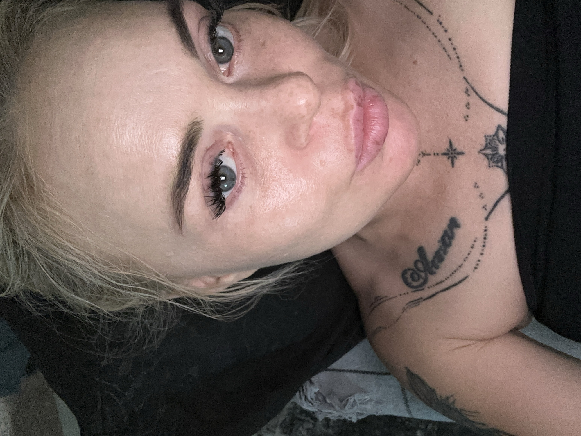 A close-up, top-down portrait depicts an adult female with blonde, disheveled hair and light-toned skin, exhibiting visible freckles on the nose and cheeks. Her blue-grey eyes are open, with prominent eyelashes and dark, defined eyebrows. Her pink lips are full and slightly pursed, creating a 'duck face' expression. Her direct gaze is towards the camera. She is wearing a black strapless garment, revealing her shoulders and upper chest. Multiple black tattoos are present: a cursive script reading "Annon" or "Annan" with decorative dotted lines on the left collarbone, a small four-pointed star with a dotted line extending upwards, and a stylized lotus flower design on the central chest. A partial tattoo, possibly a feather or wing design, is visible on her right shoulder and upper arm. The background is largely dark and indistinct, with a light-colored, textured, and frayed fabric visible on the left side behind her shoulder. The lighting is soft and appears to be artificial, highlighting her facial features. The specific location in the city of Unknown, Unknown, is not discernible from the image.