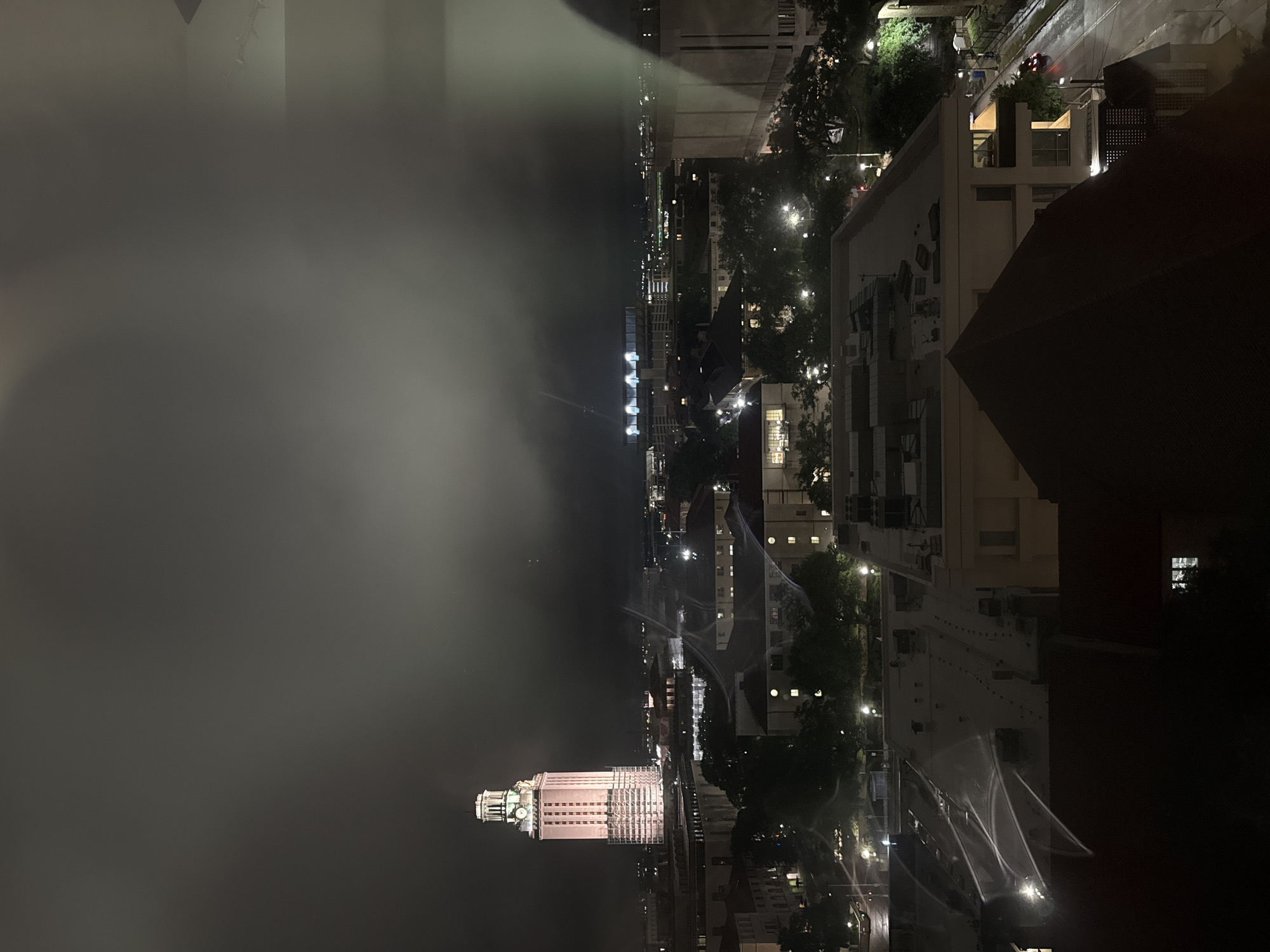 The image shows a nighttime view of the University of Texas Tower in Austin, Texas. The tower is illuminated and partially obscured by fog or clouds. Surrounding buildings and streets are visible, also illuminated by streetlights. There are no people clearly visible in the image. The overall environment is dark and foggy.