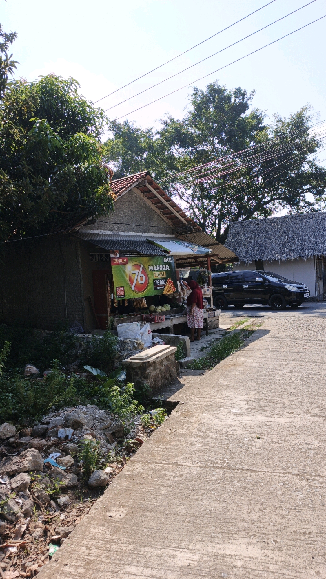 A street-level, medium shot captures a roadside stall in Desa Batukaras, Indonesia, on a sunny day. The stall, constructed from concrete and corrugated metal, is situated next to a paved walkway and a dirt path littered with rocks and debris. A prominent banner advertising "BARU MANGGA ROYAL" hangs at the front of the stall, indicating a fruit vendor. A woman in a red headscarf and a patterned skirt is interacting with the vendor, who is partially visible behind a display of produce. Several large white sacks and various fruits, including cabbages, are visible on wooden shelves. Behind the stall, a black minivan is parked on the road. Lush green trees and a building with a thatched roof are visible in the background, and power lines are strung across the sky.